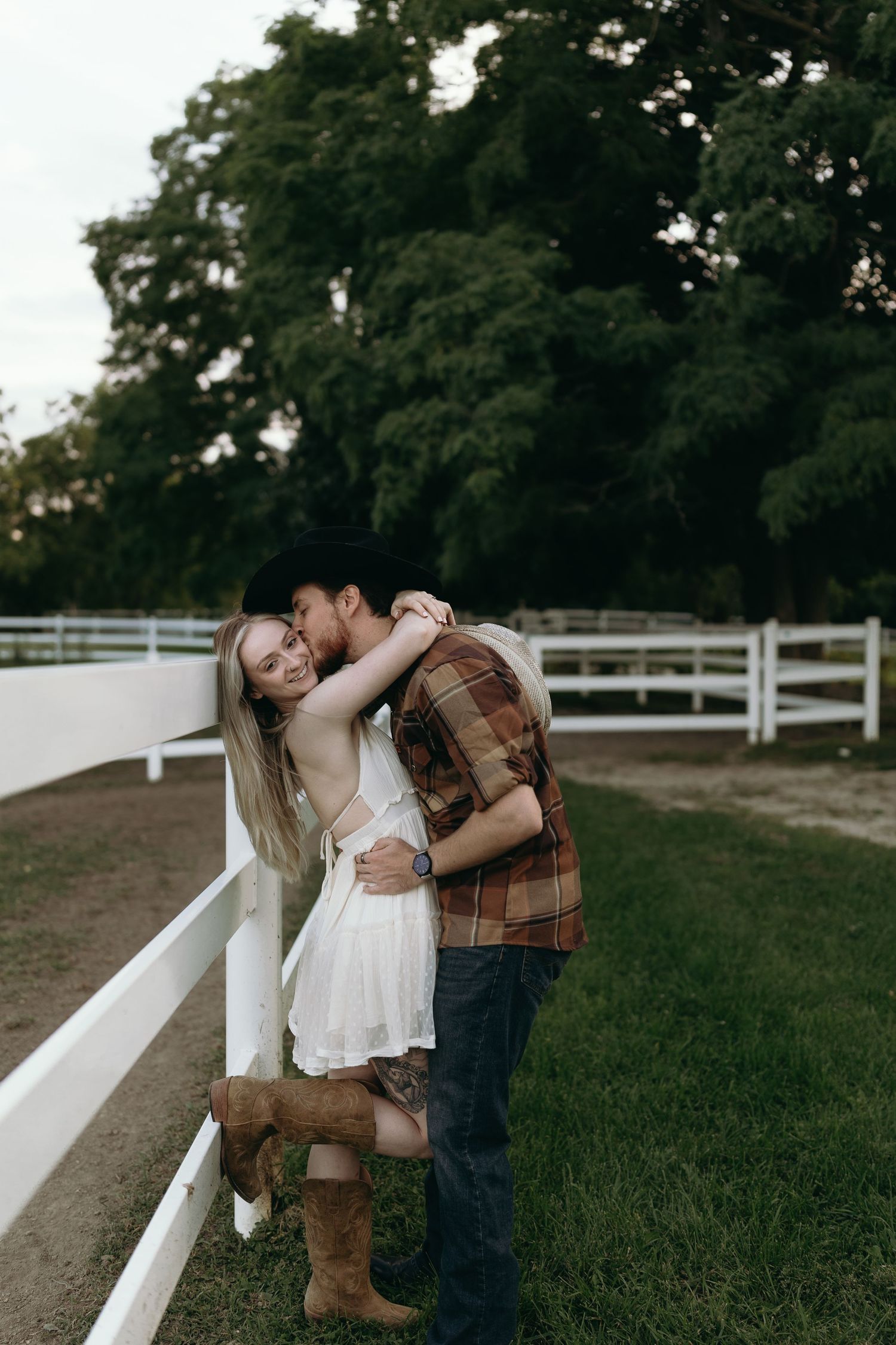 A romantic couple embraces by a white fence at sunset on a grassy hillside.