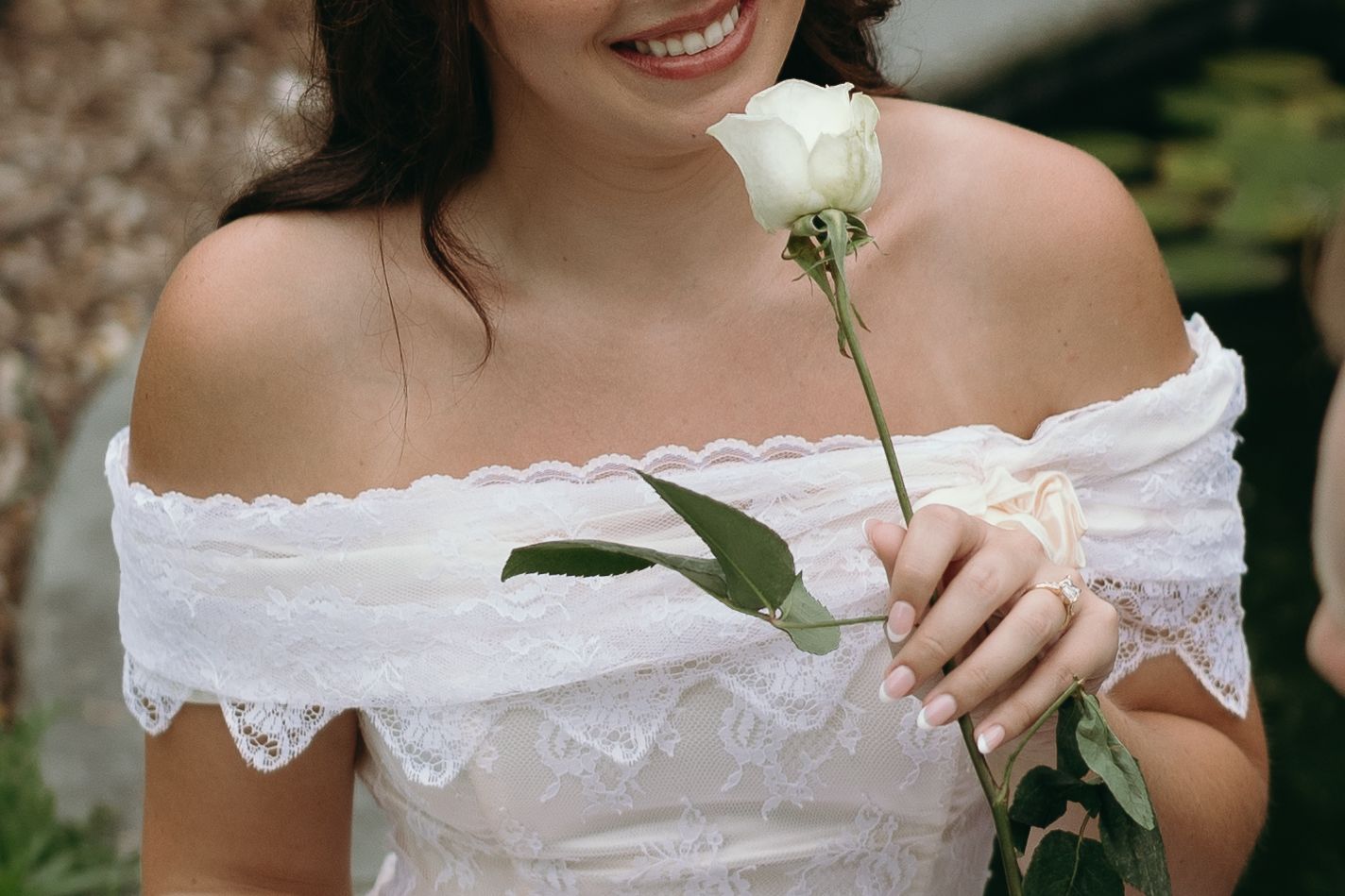 A person in a white lace dress holding a single white rose flower while smiling outdoors.