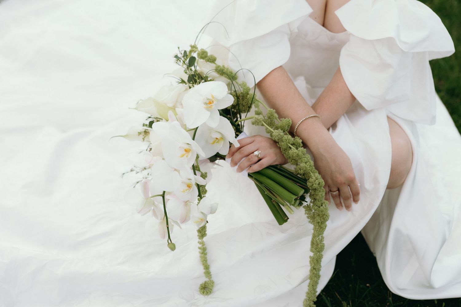 Bride in white gown holding elegant cascading bouquet of white orchids and green accents while seated outdoors.