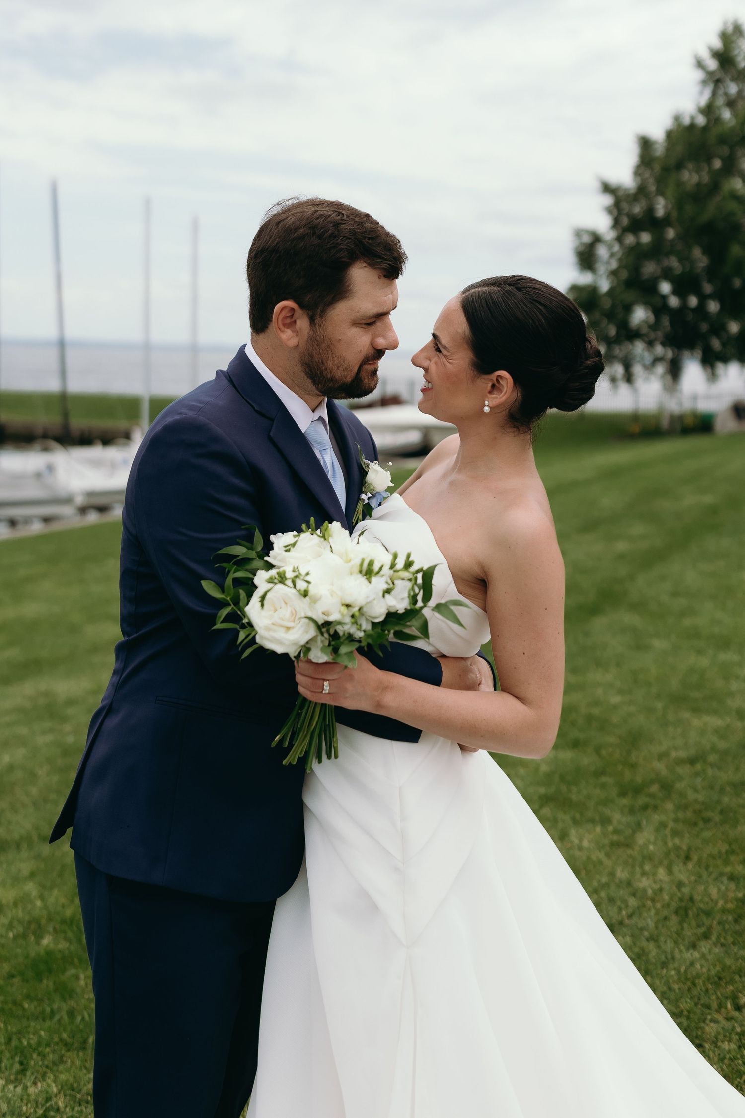 A romantic wedding couple embraces on a grassy lawn overlooking a scenic waterfront view.