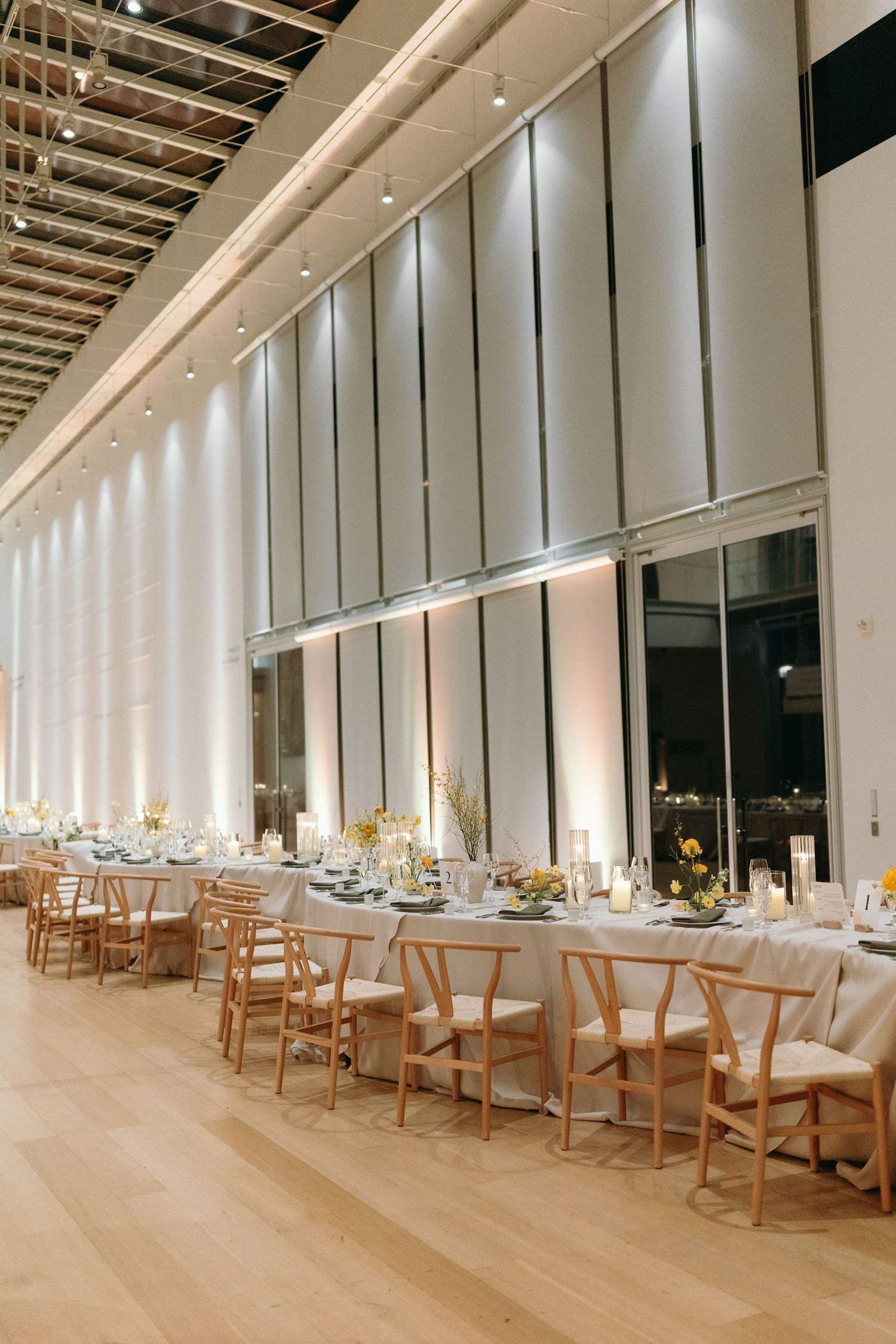 An elegant wedding reception setup with wooden chairs and white linens in a modern high-ceiling venue.