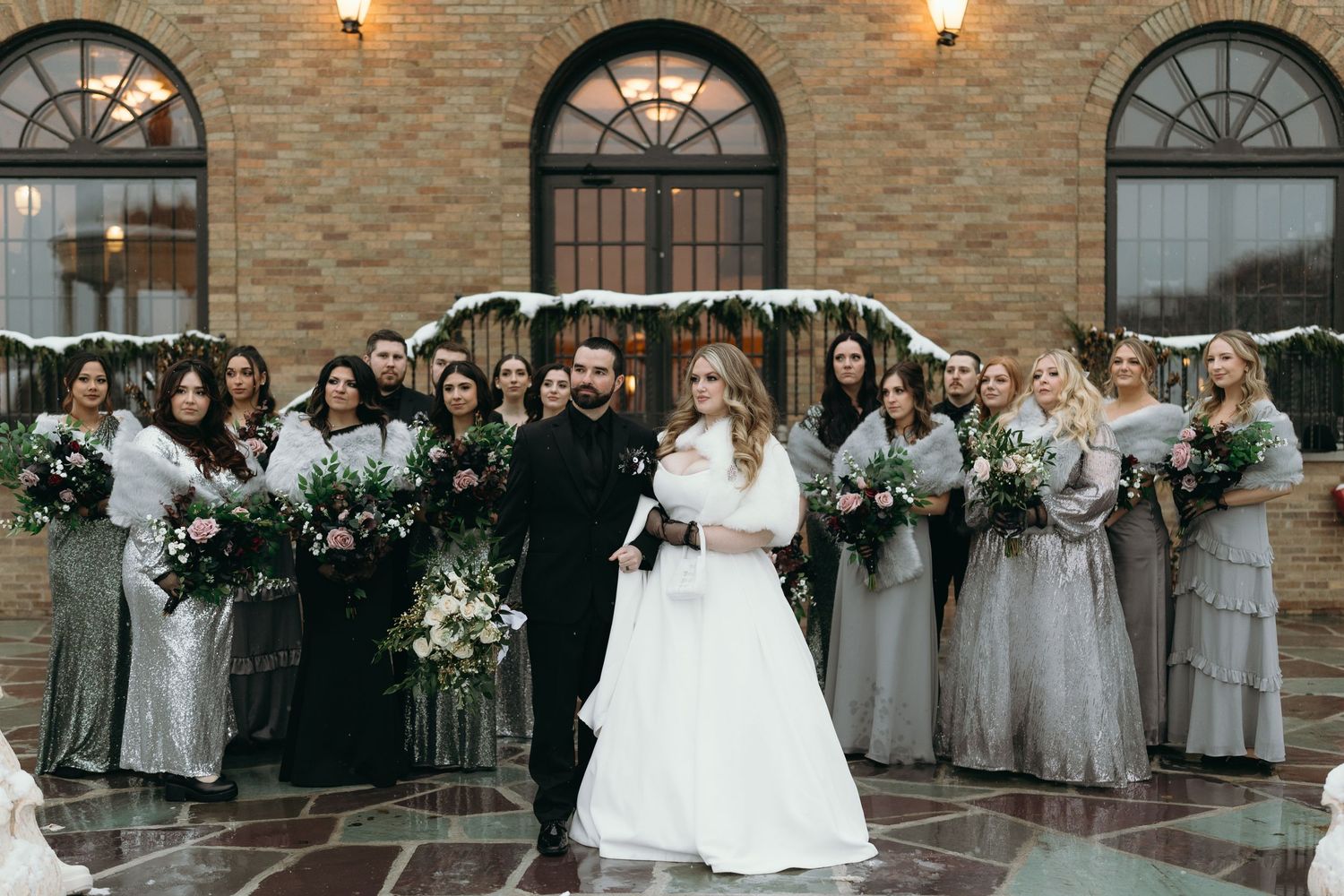 Wedding party in gray dresses and black suits pose together under brick archway.