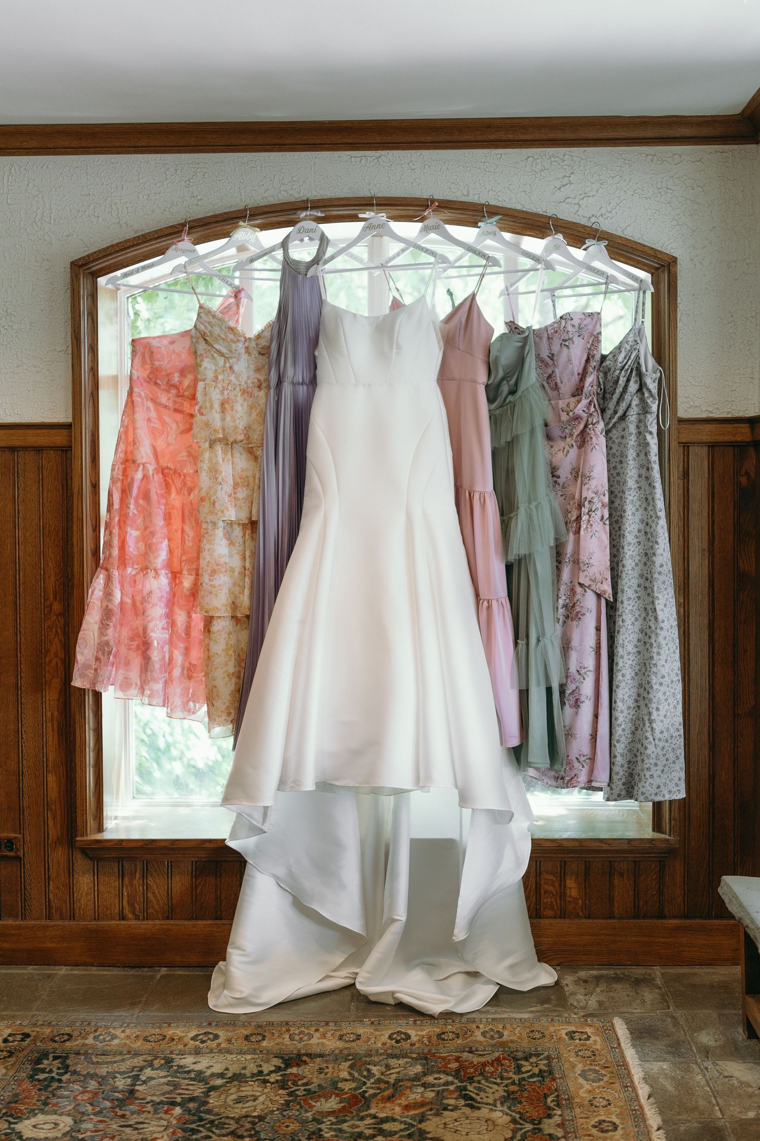 Elegant bridesmaid dresses in coral, lavender and white hang against a wood-paneled wall near a curved window.