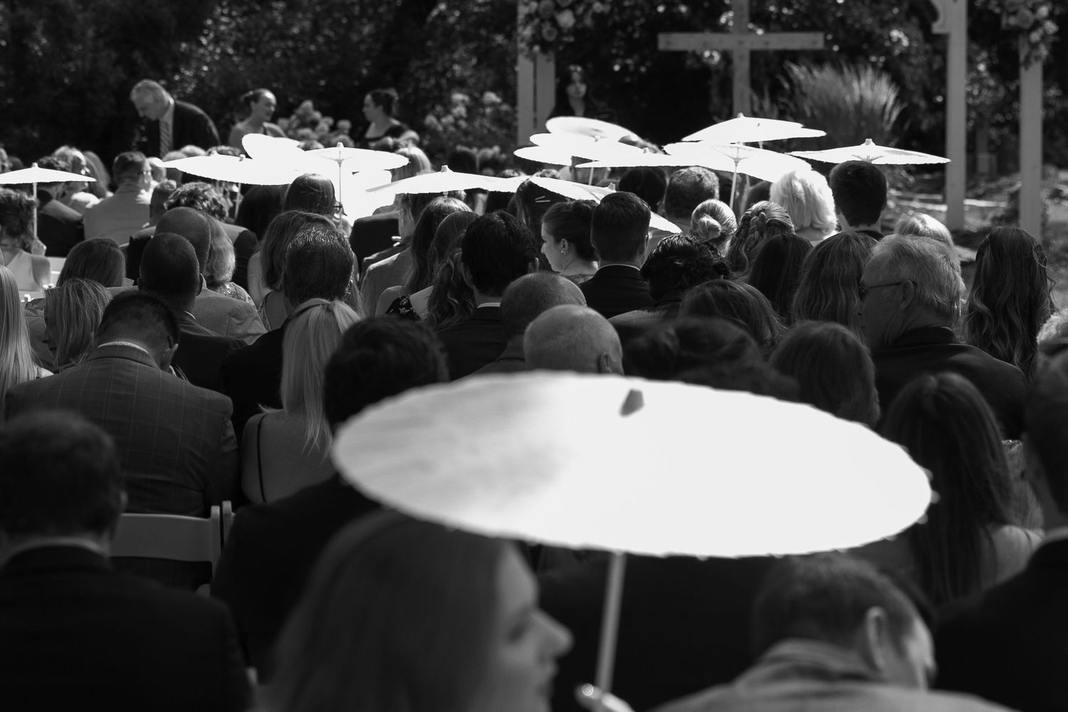 Black and white photo of wedding guests seated outdoors, holding white parasols during an elegant garden ceremony.