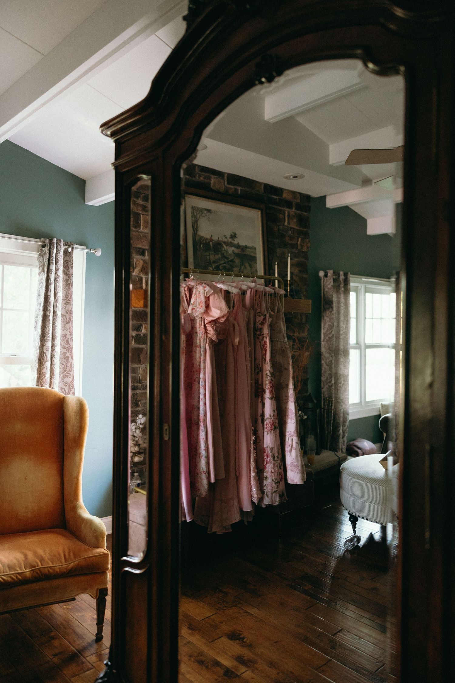 Elegant vintage dressing room with dark wood armoire mirror reflecting pink dresses on rack, brick wall, and antique wingback chair.