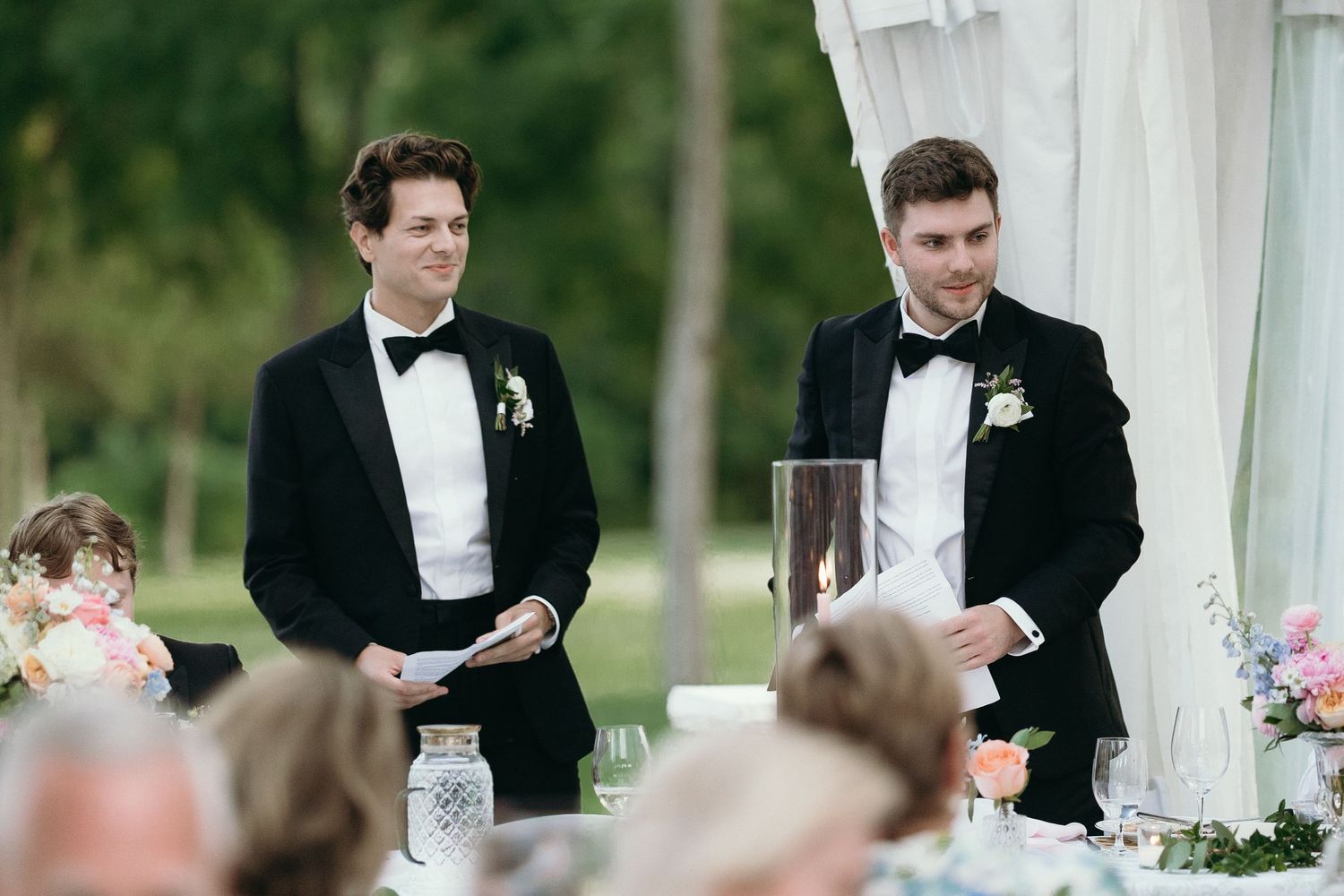 Two groomsmen in black tuxedos deliver speeches at an elegant outdoor wedding reception.