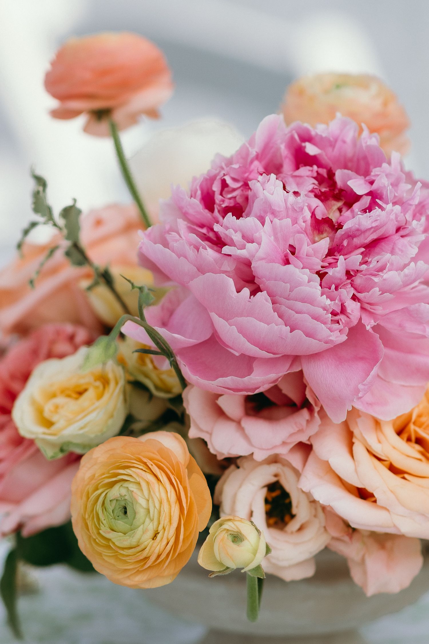 Elegant floral arrangement featuring a large pink peony surrounded by peach ranunculus and roses in soft pastel tones.