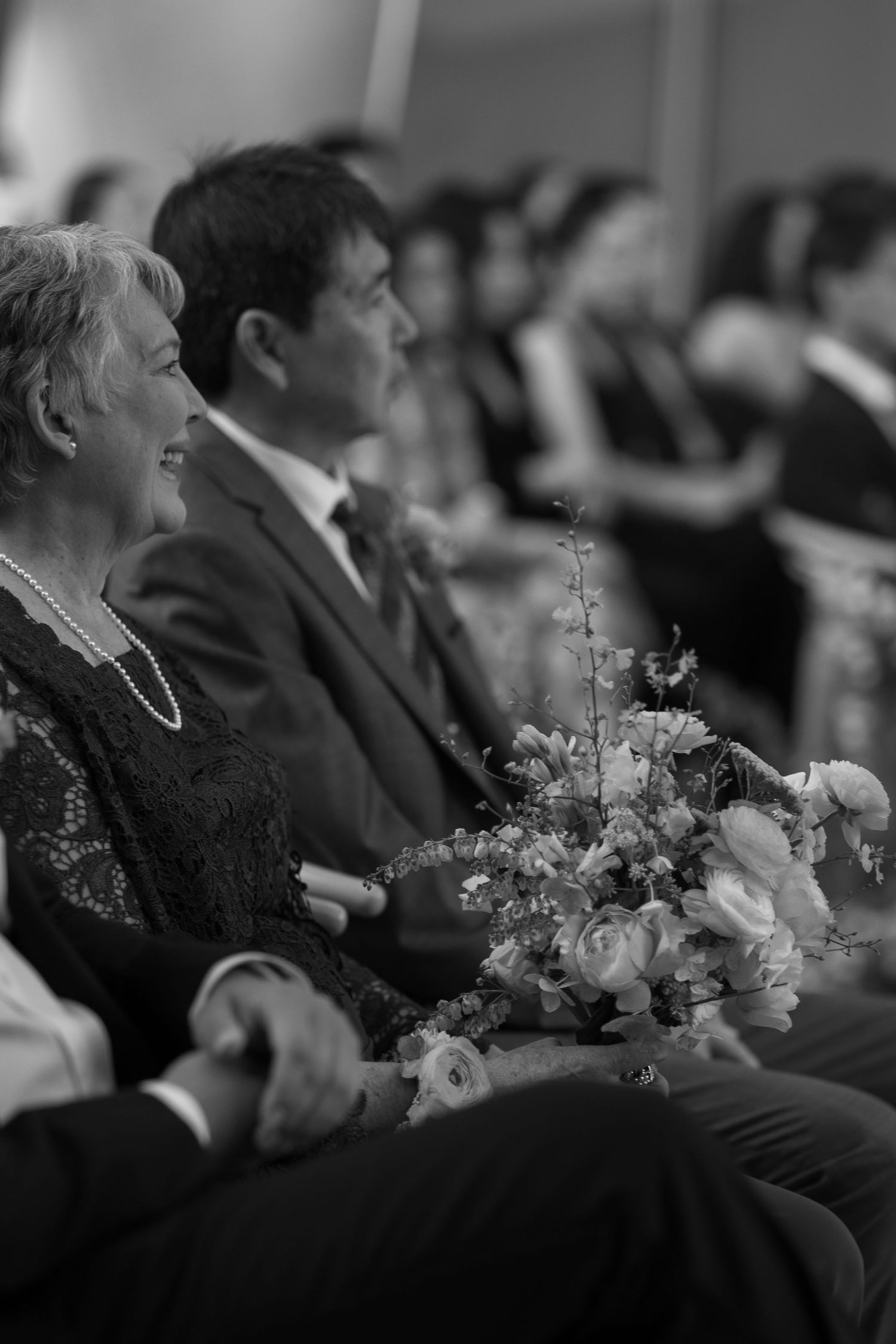 Wedding guests watch ceremony with expressions of joy as floral arrangements adorn the seating area.