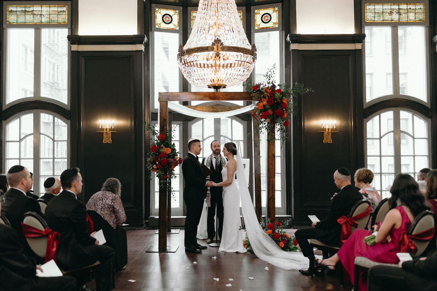 Wedding ceremony in a grand room with dark wood panels and ornate lighting fixtures.