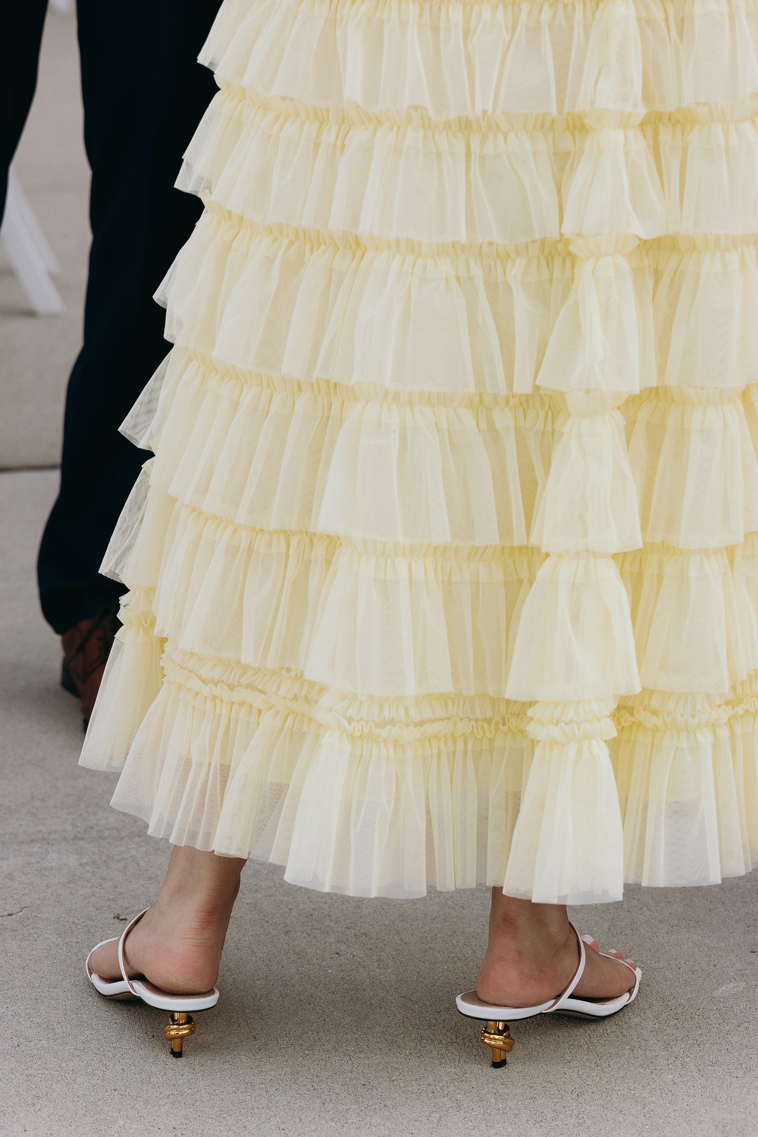 Close-up view of layered cream tulle ruffles on a dress with white kitten heel mules peeking out at the bottom. Cocktail hour inspo