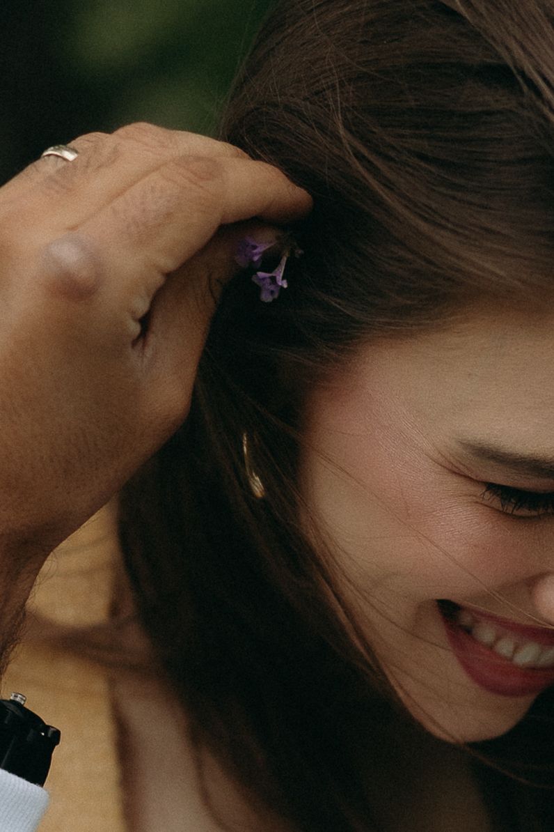 A man places a small purple flower in a smiling woman's brown hair, wearing a ring and black watch.