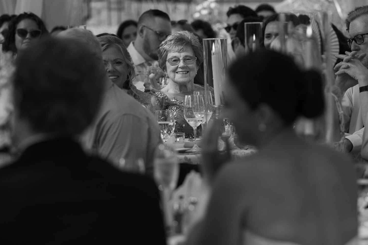 Black and white wedding reception photo of smiling elderly woman surrounded by guests at elegantly set dinner table.