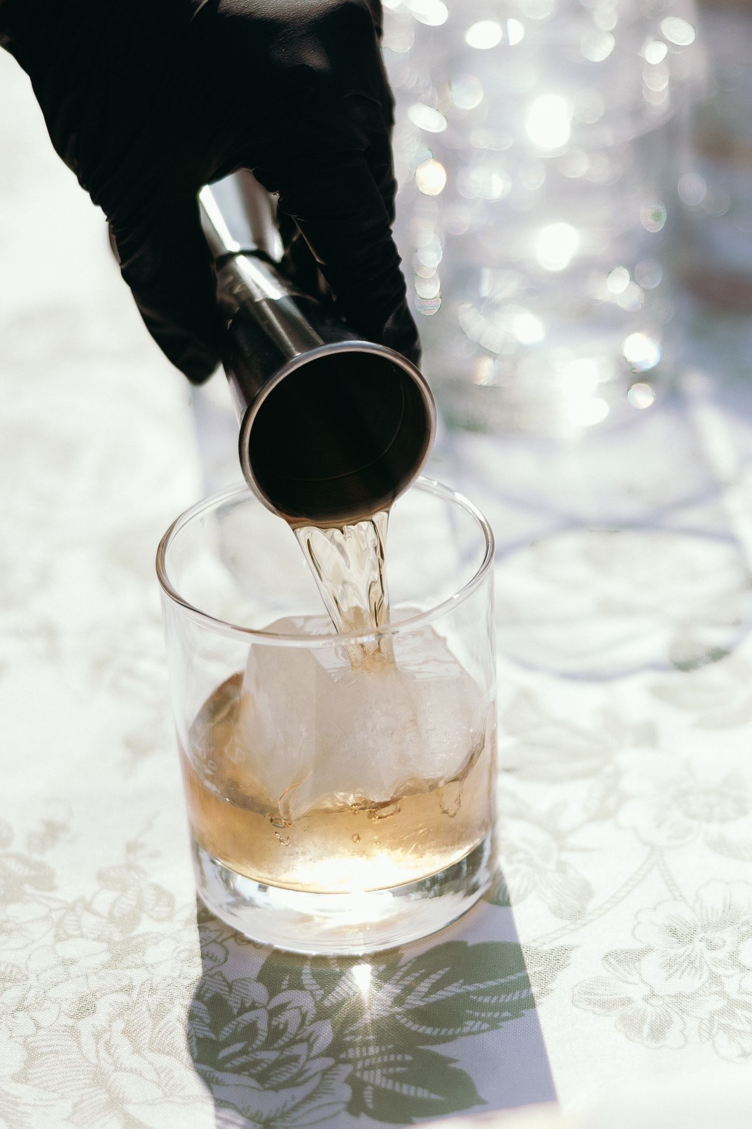 Bartender in black gloves pouring whiskey from a jigger into a rocks glass with ice.