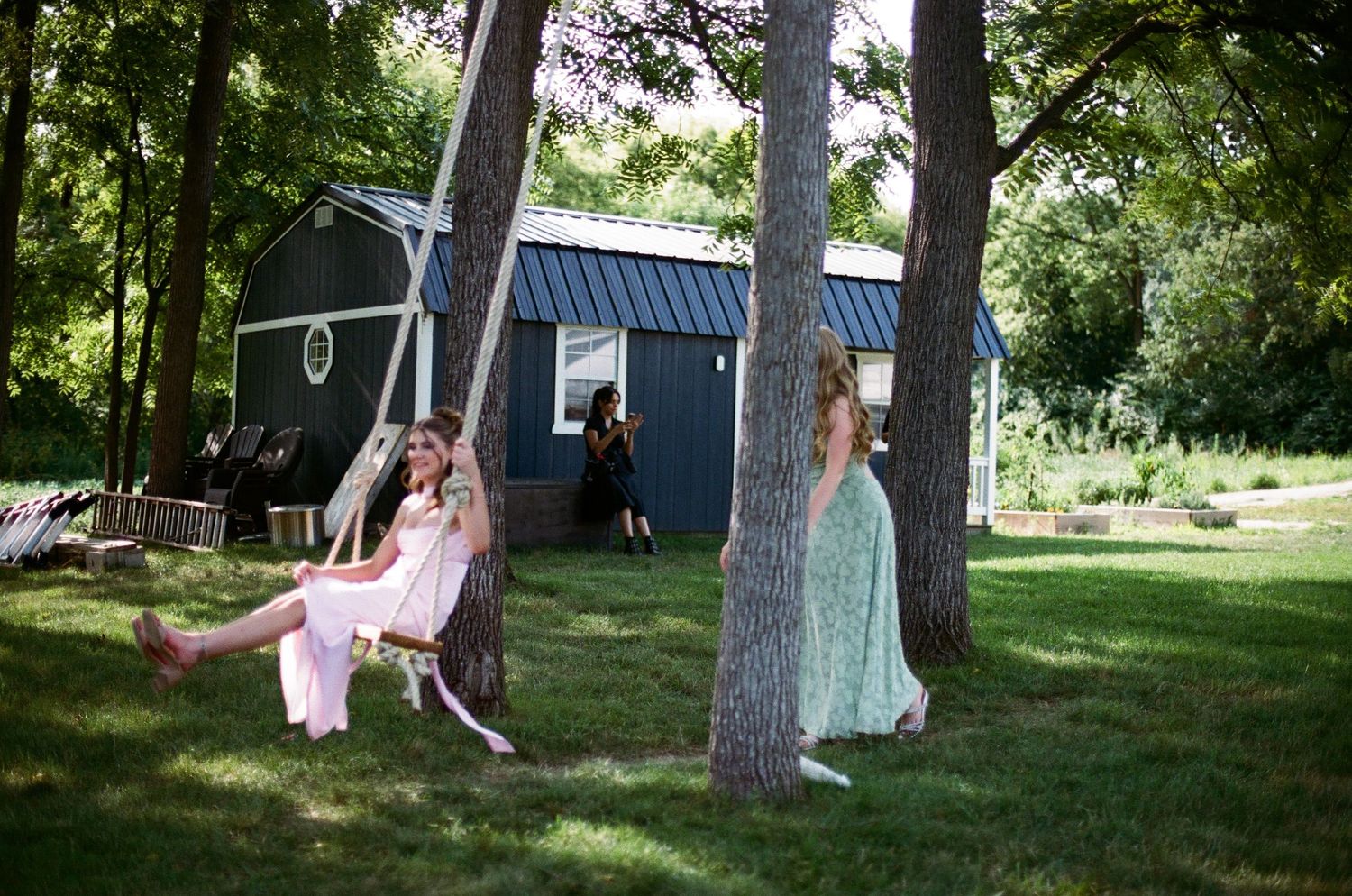 Two girls in formal dresses enjoy a tree swing outdoors near a dark barn on a sunny day.