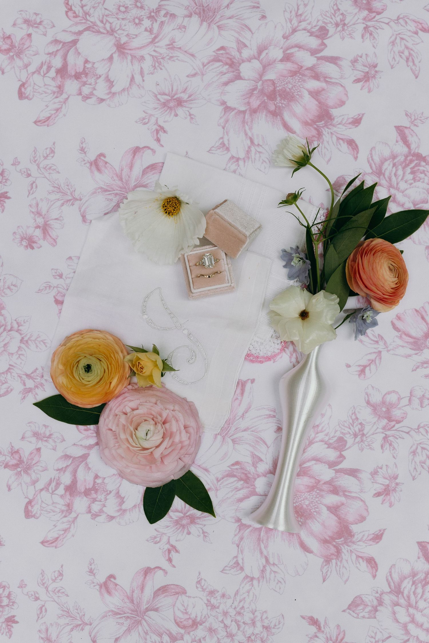 Romantic flat lay of pink ranunculus flowers, white blooms, a ring box, and a silver vase on floral toile fabric.