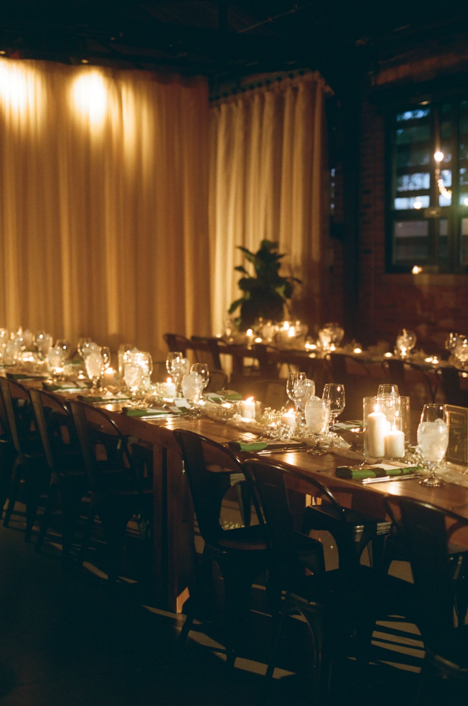 Elegant candlelit wedding reception with long wooden tables, glassware, and cream drapery in a warm, intimate venue.