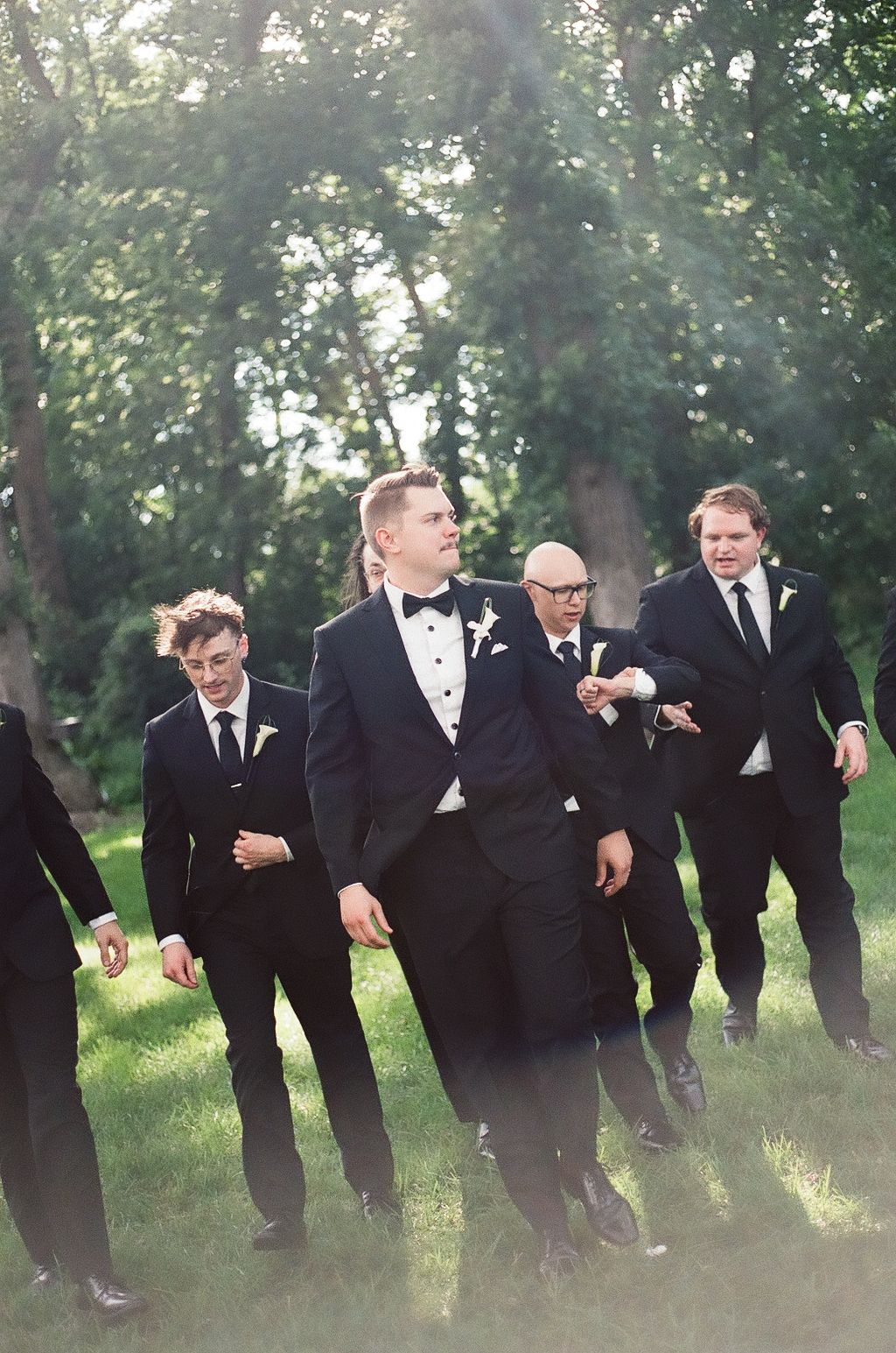 Groomsmen in black tuxedos with bow ties and boutonnieres walking outdoors on green grass at a wedding.