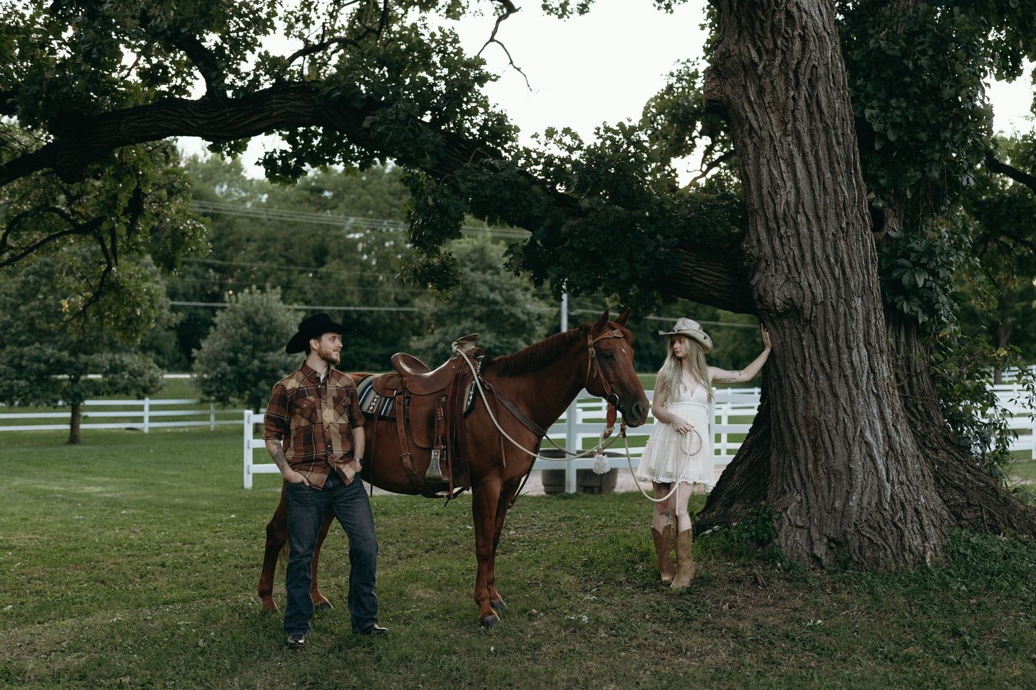 People riding horses along a country trail near large trees and white fencing on a summer day.
