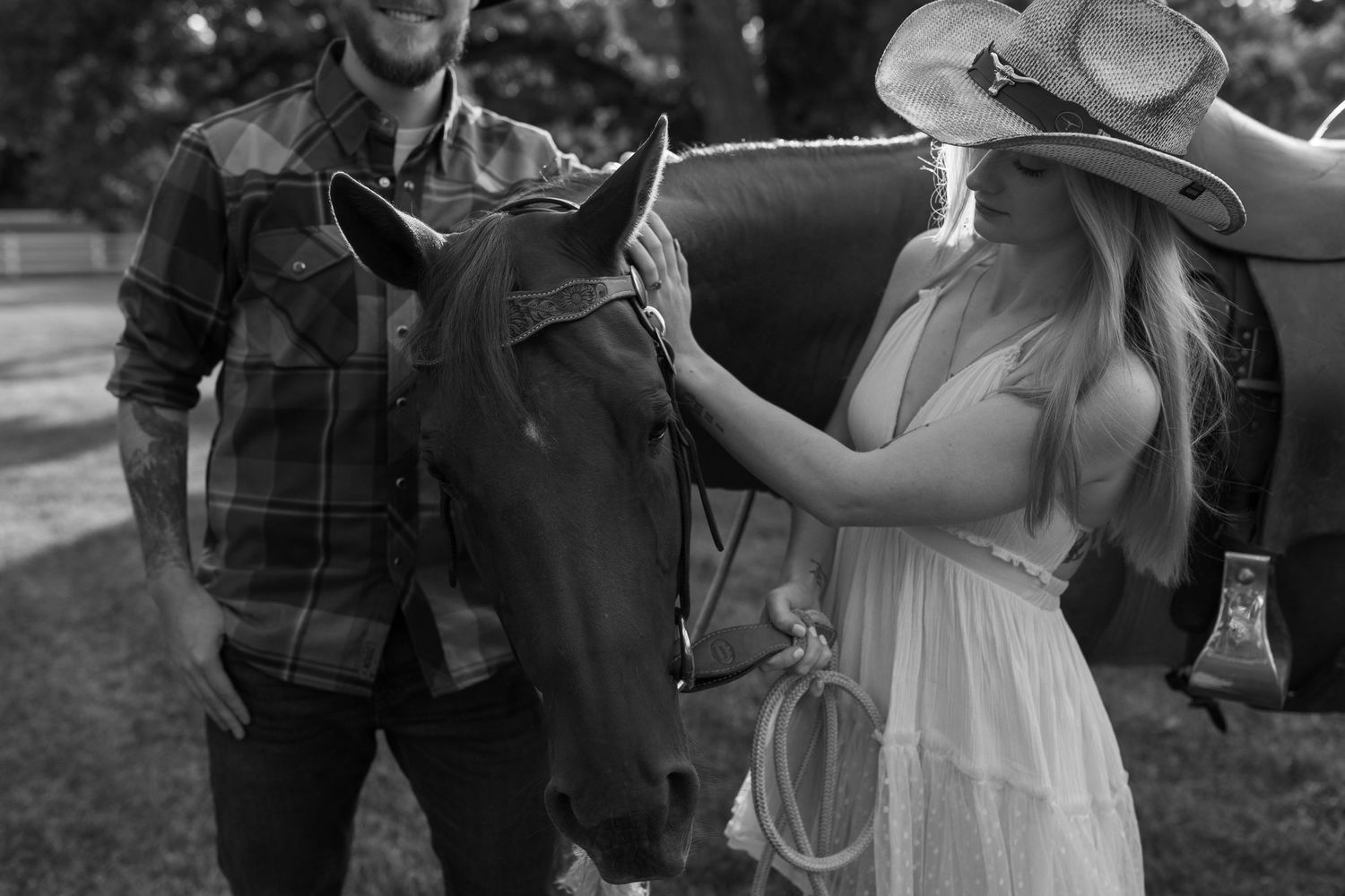 A couple in casual western attire stands with a dark horse between them in a black and white outdoor setting.