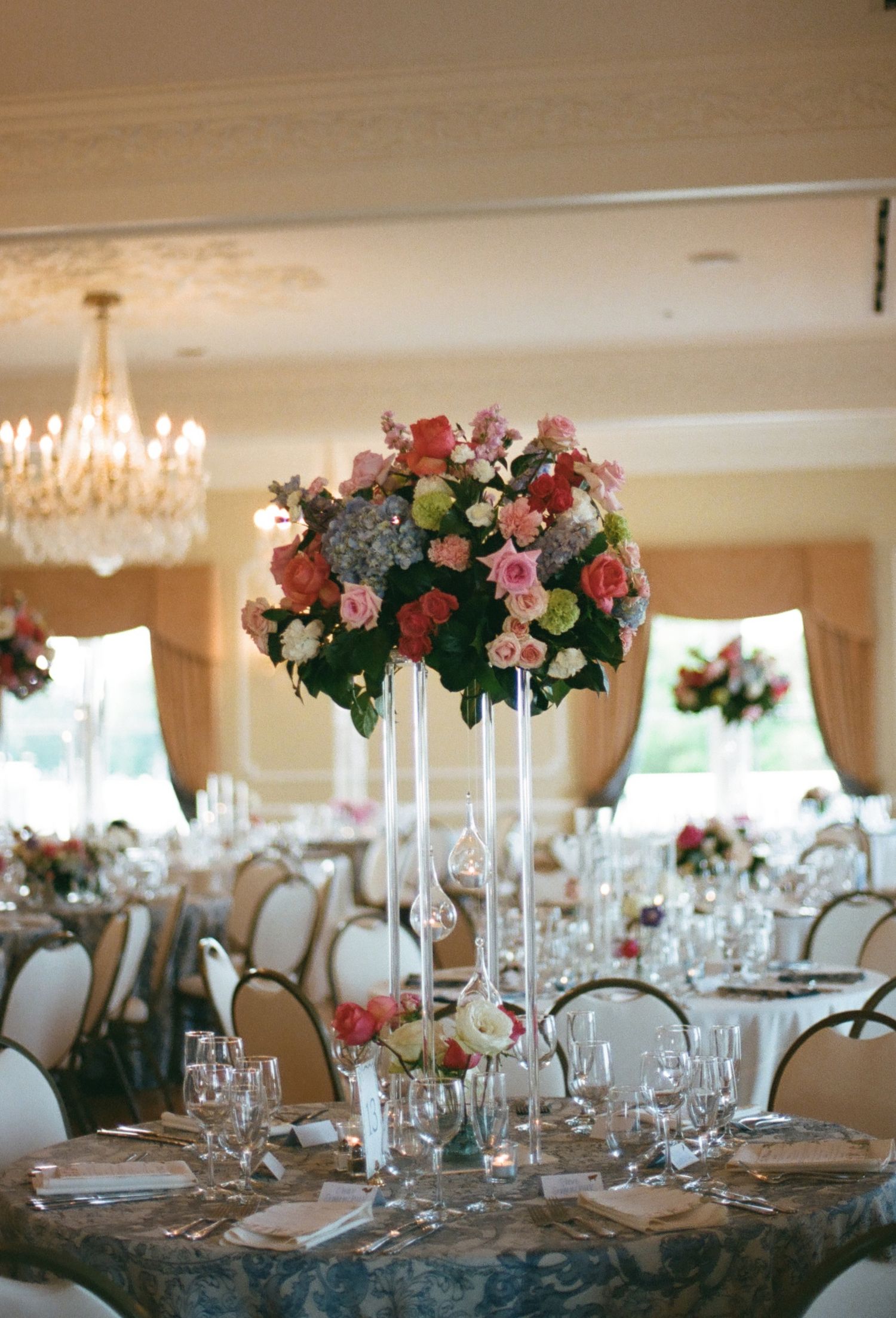 Elegant wedding reception centerpiece with pink roses and hydrangeas on tall glass stand, crystal chandelier ballroom.
