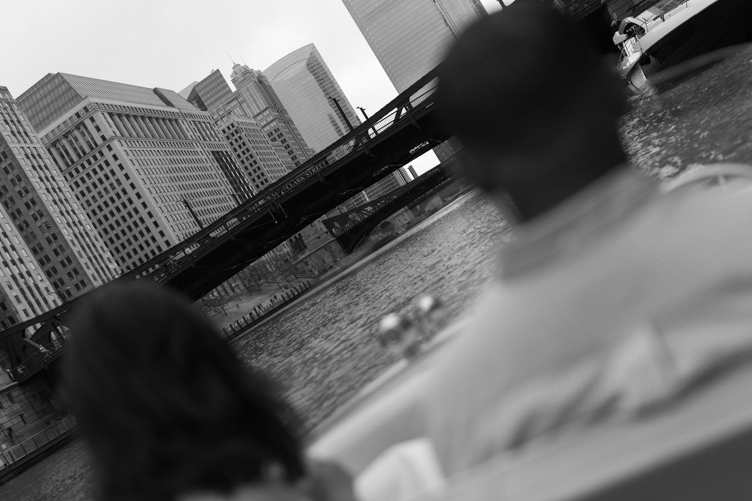 Black and white photo of two people overlooking a city river with a bridge and skyscrapers in the background.