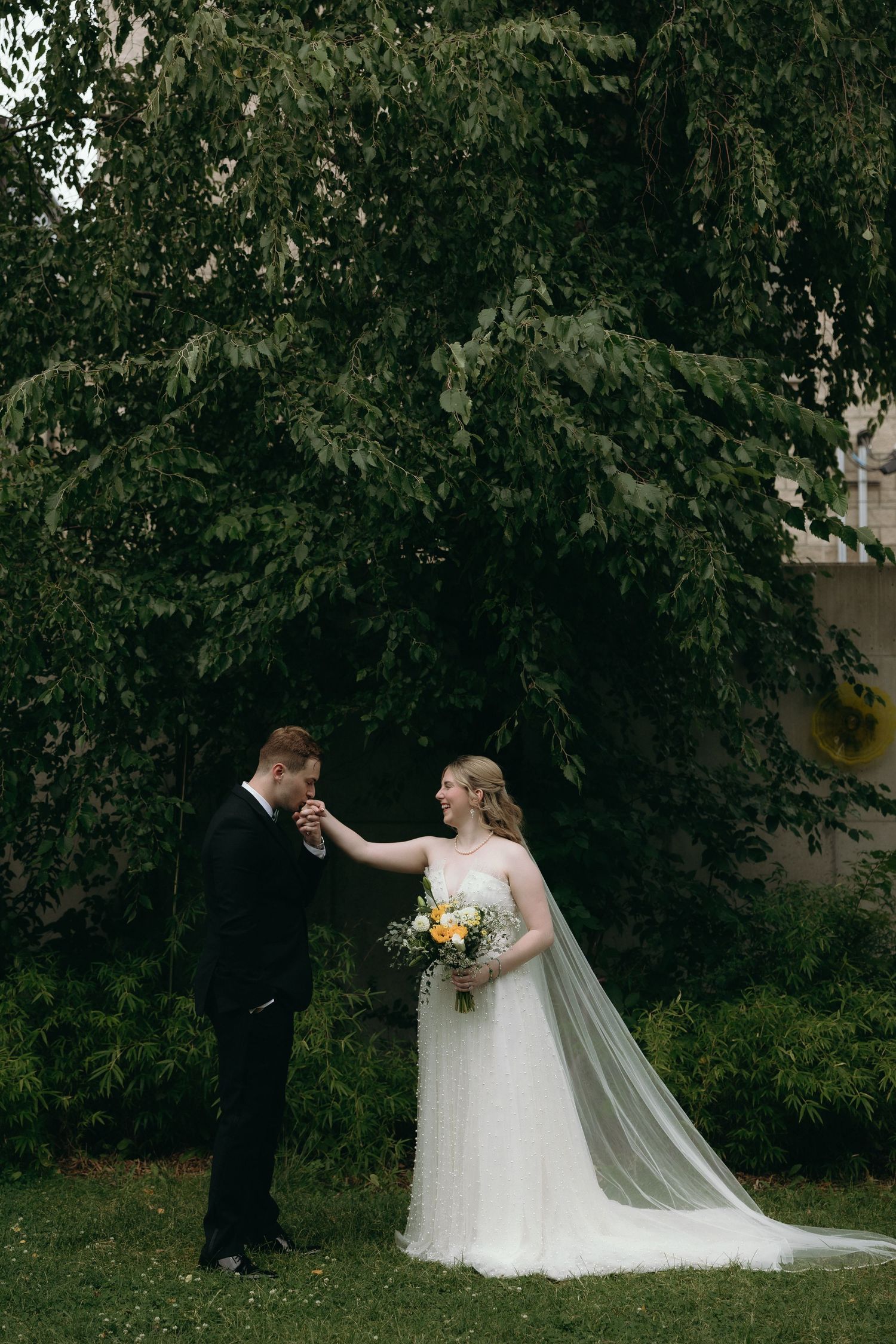Groom in black suit kisses bride's hand outdoors, bride wearing white gown with veil holding yellow floral bouquet.