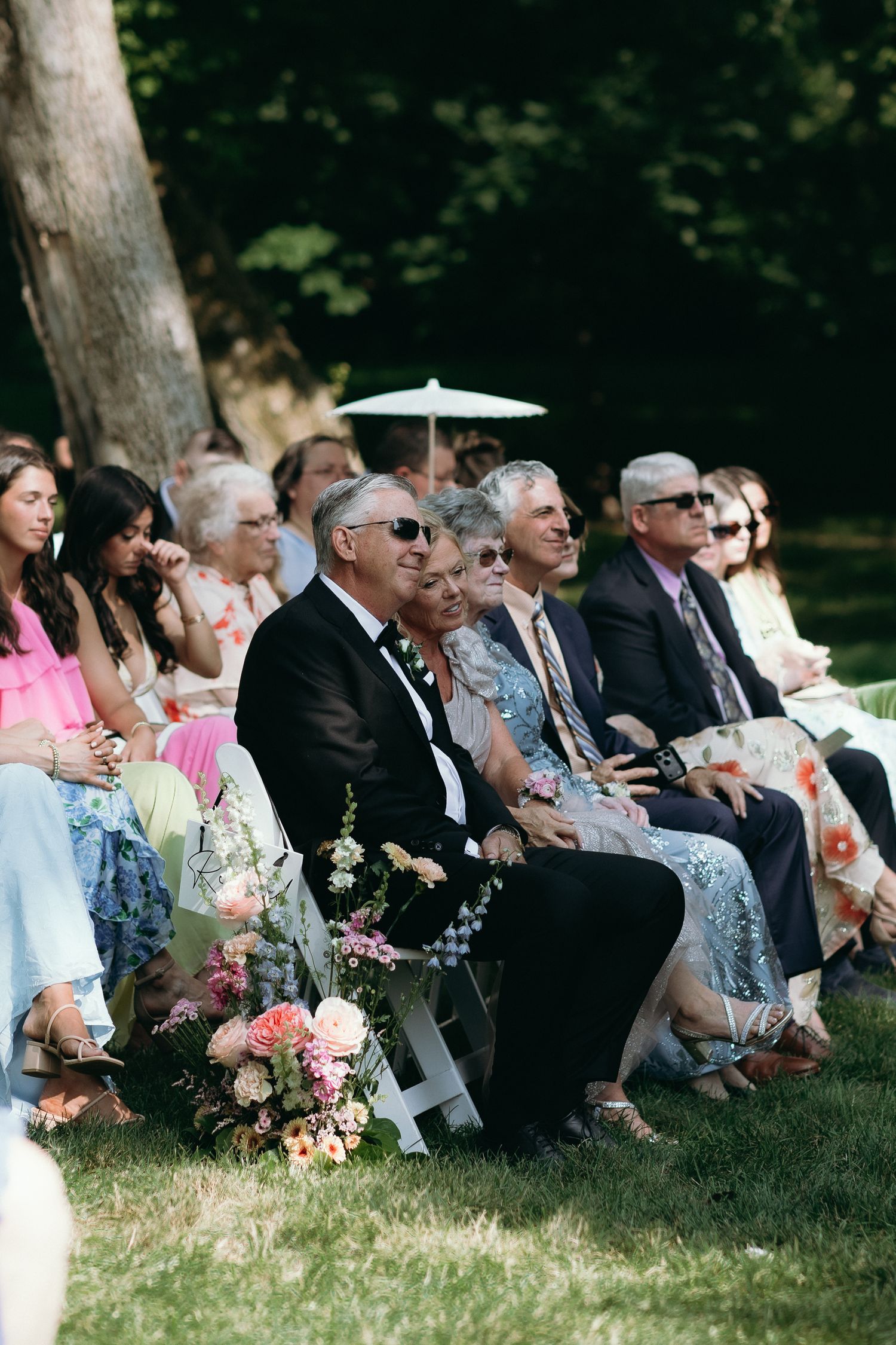 Wedding guests seated on white chairs outdoors watching ceremony under shaded trees on a bright summer day.