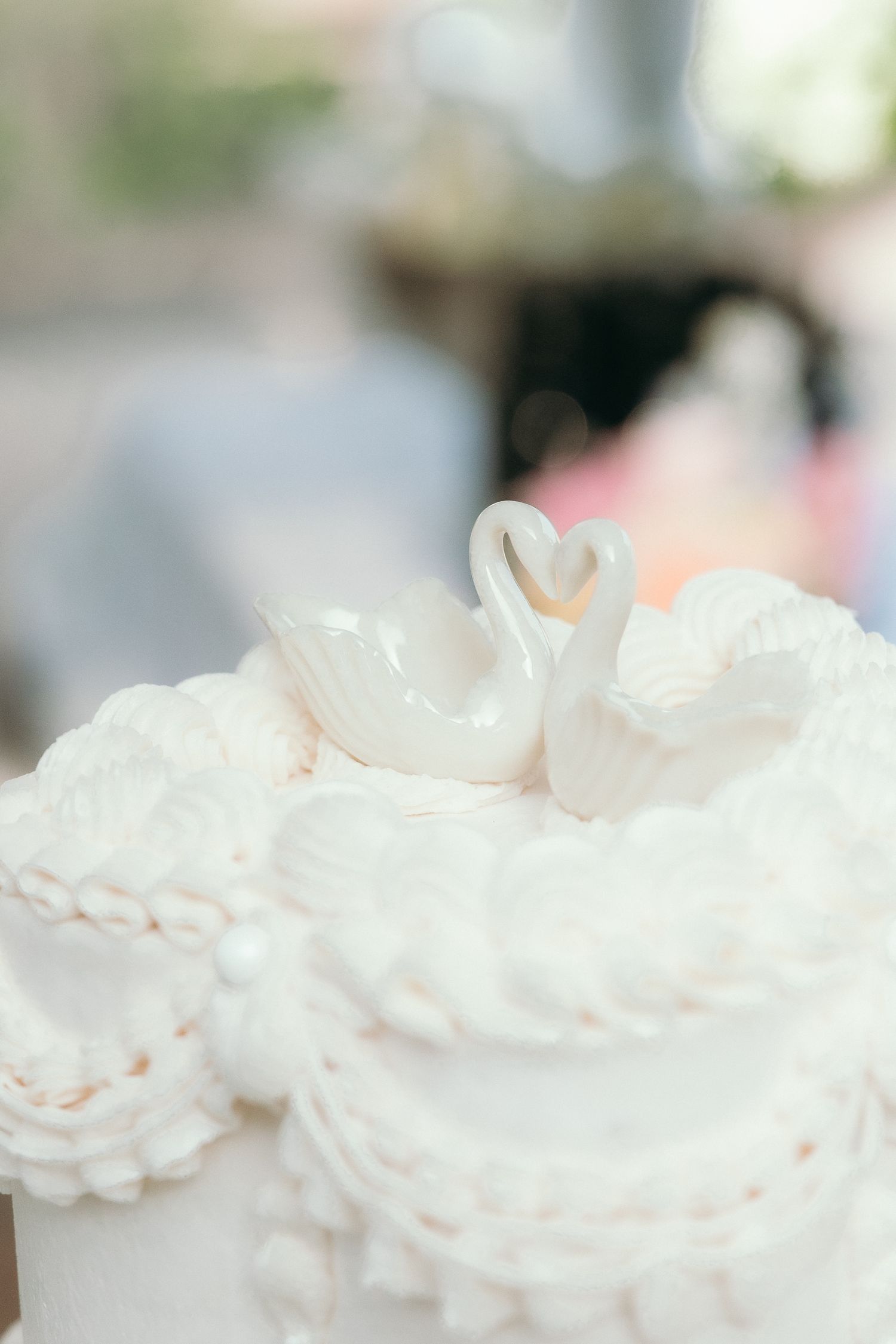 White wedding cake topped with two swan figurines forming a heart shape, decorated with intricate white frosting details.