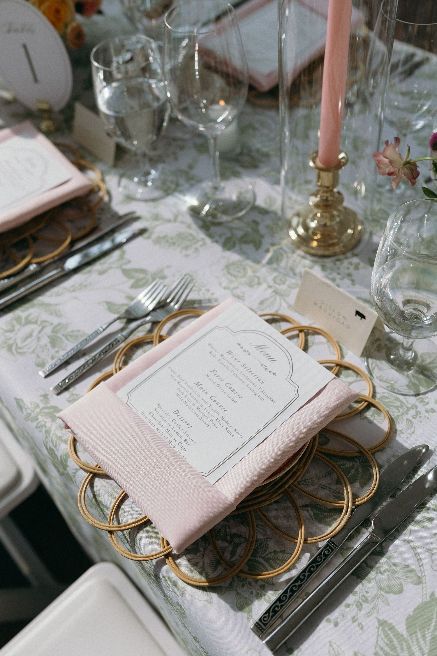 Elegant wedding table setting with gold charger plates, pink napkins, menu cards, and floral tablecloth.