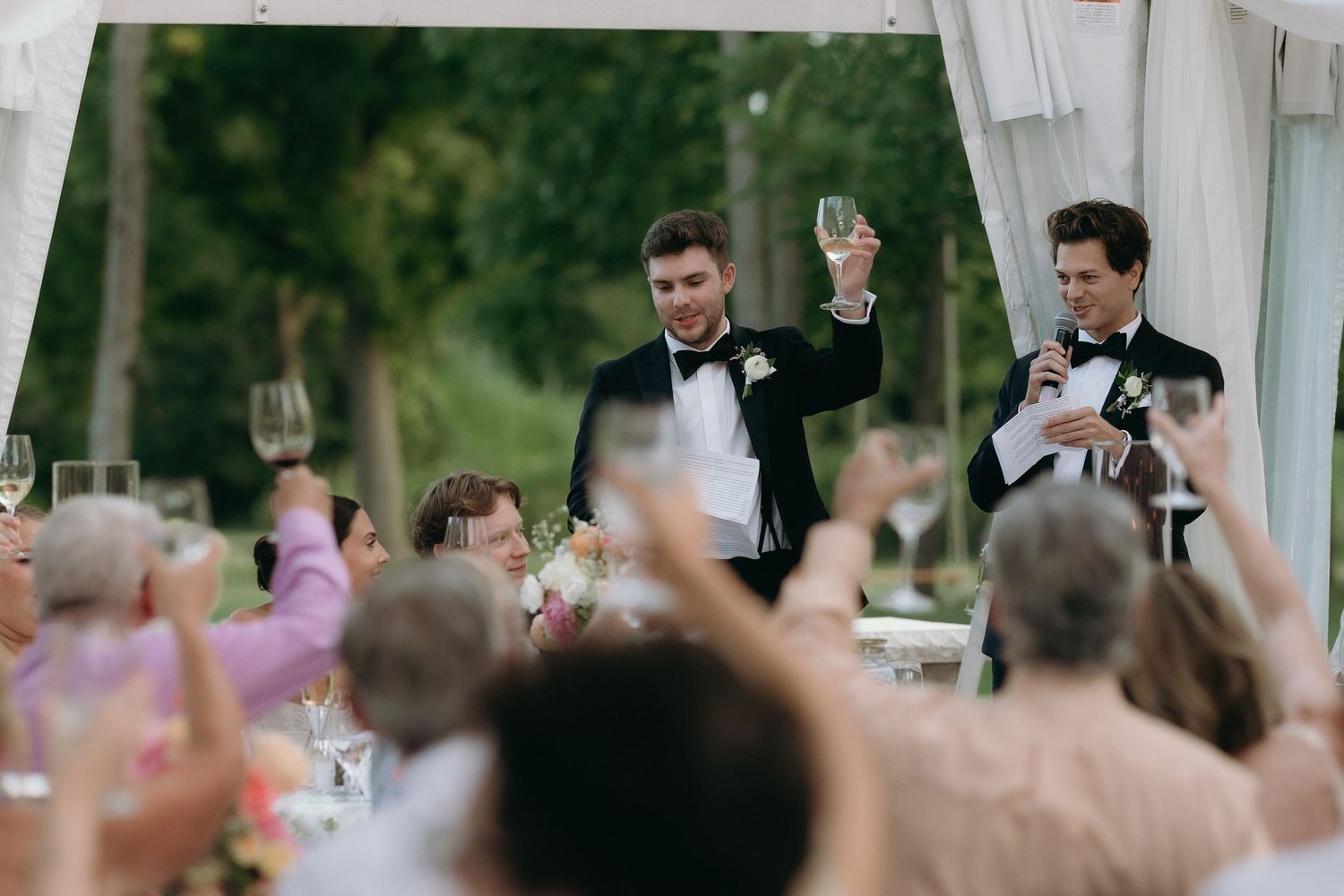 Two grooms in tuxedos raising champagne glasses for wedding toast with cheering guests.