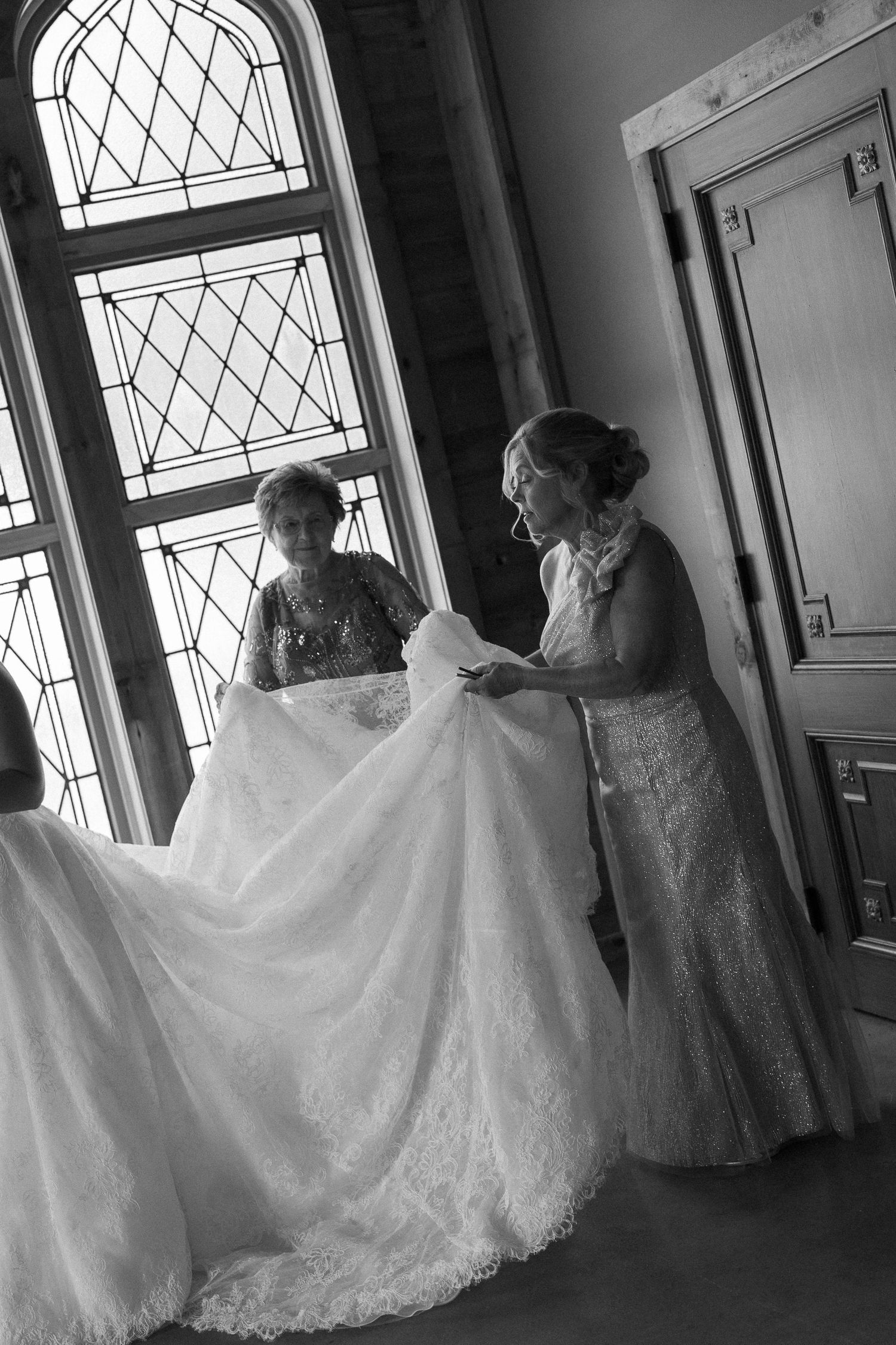 Black and white photo of two women arranging a white lace wedding gown near stained glass windows.
