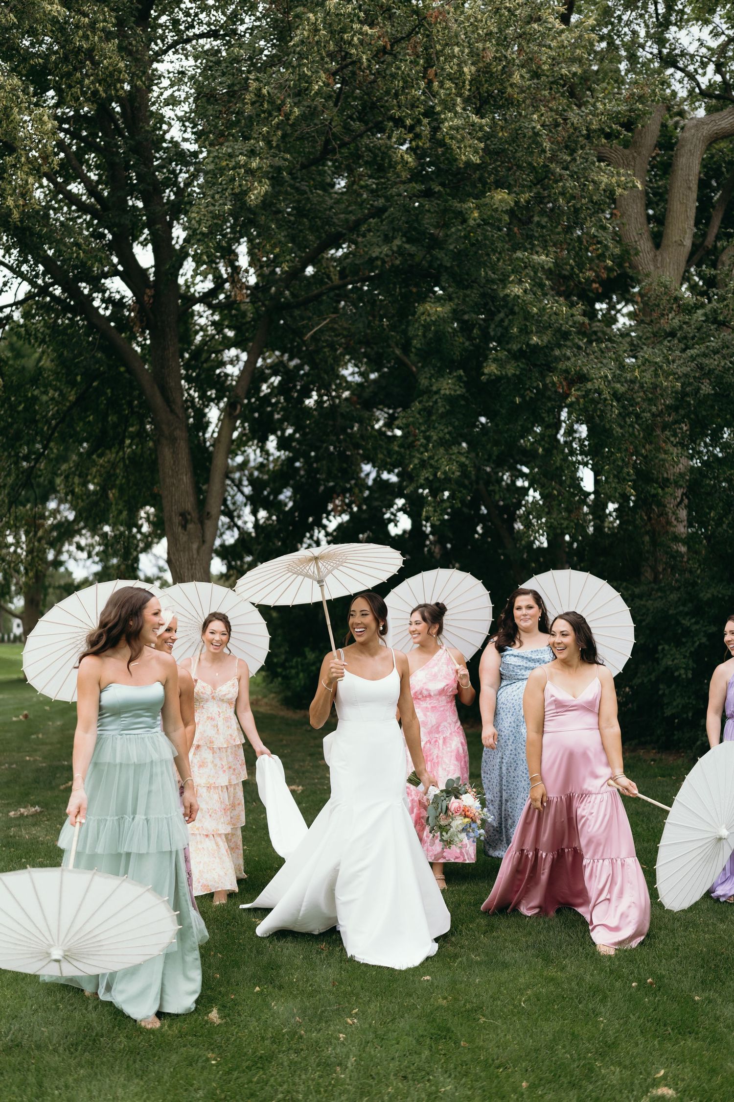 Bridal party with white parasols posing on grass under trees in summer.
