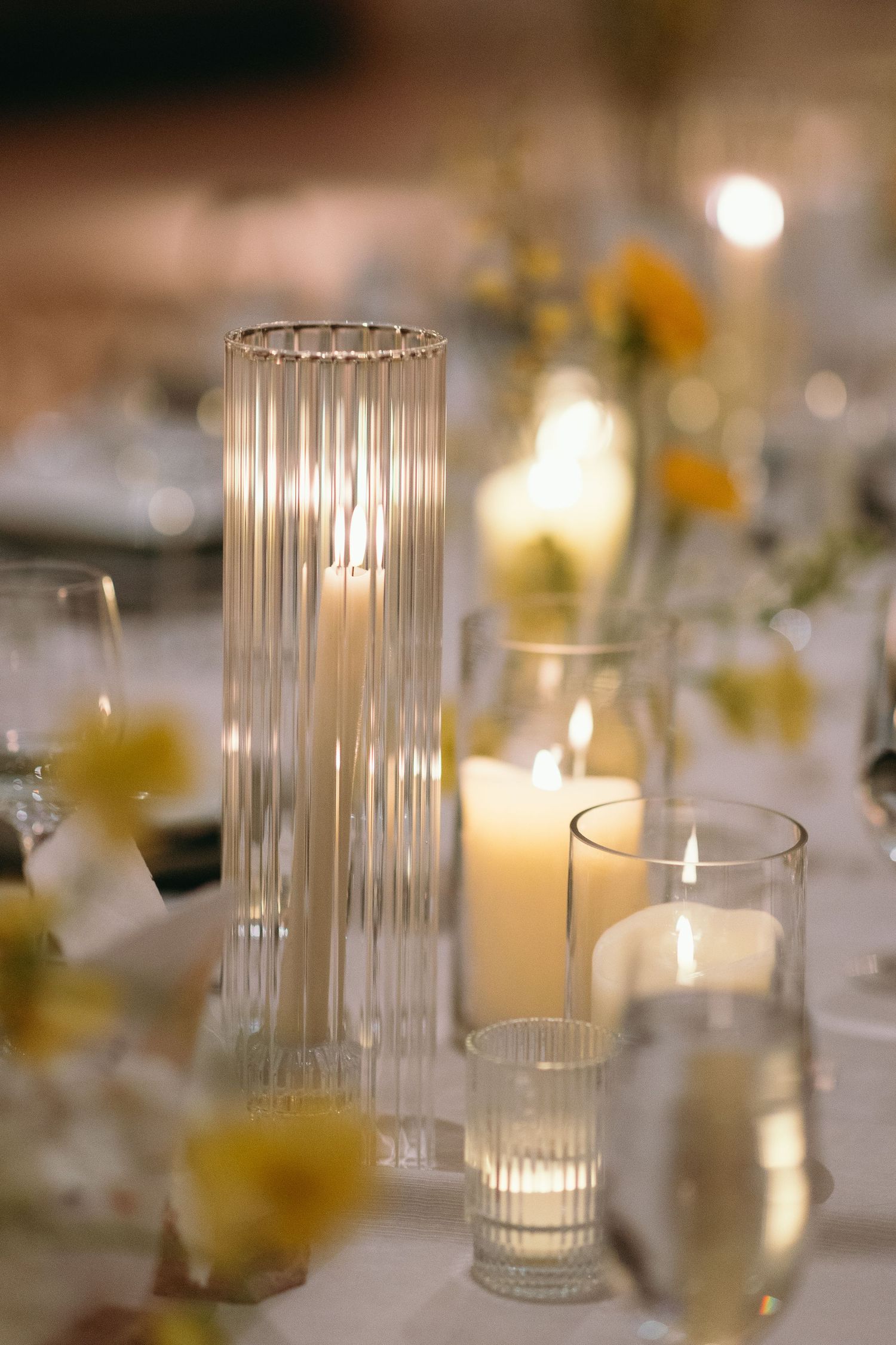 Glowing candles in ribbed glass holders with yellow flowers create romantic table ambiance.