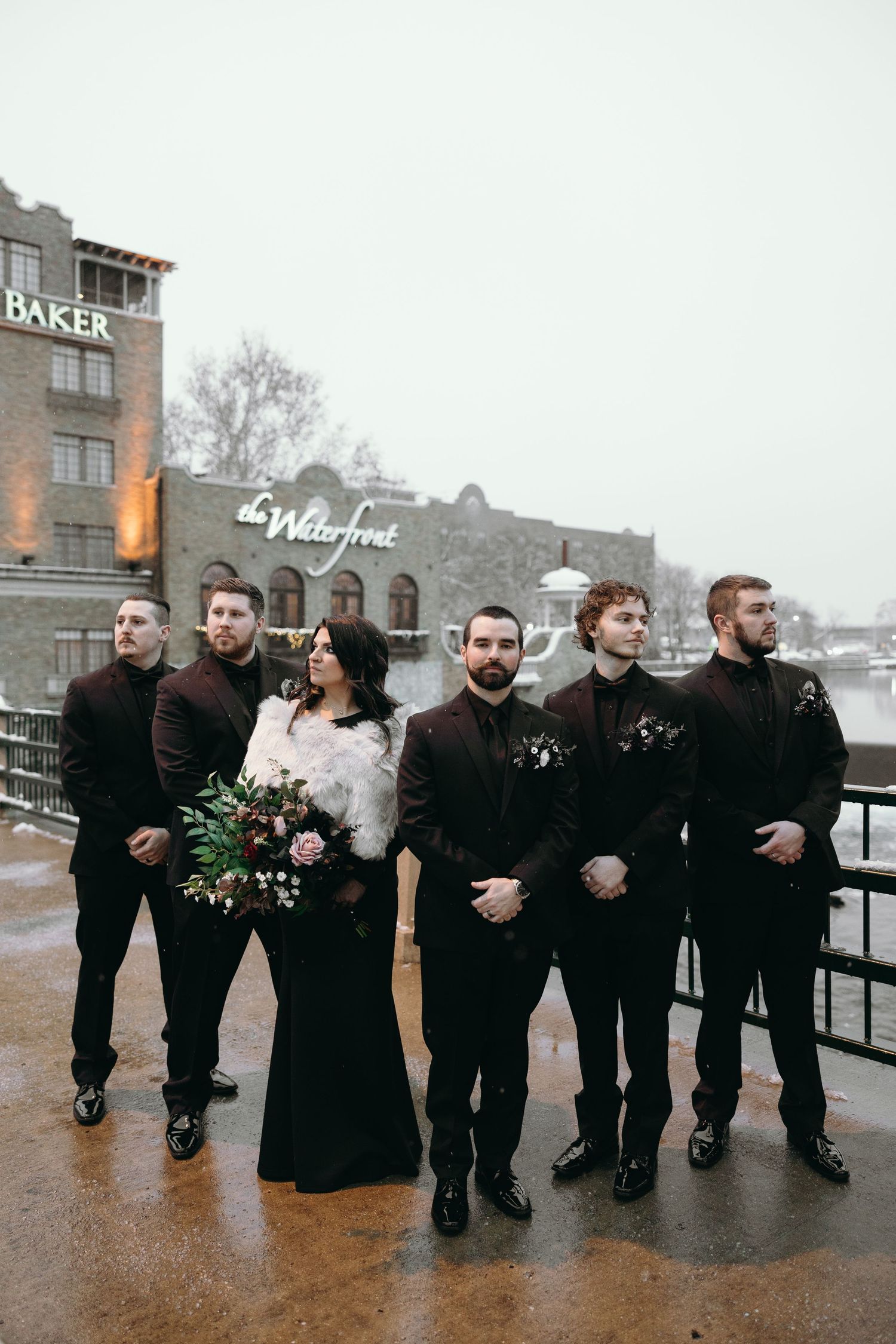 Wedding party in black attire poses outside historic brick building on rainy winter day.