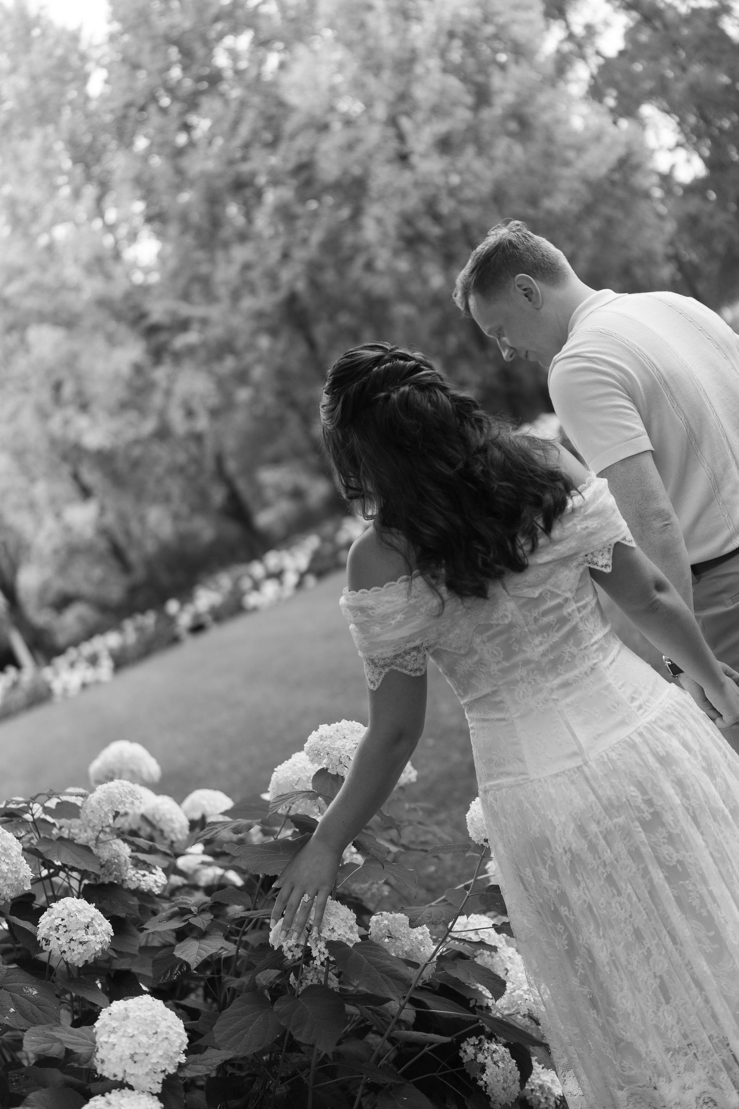 Black and white photo of someone in a white dress picking hydrangeas in a garden setting.