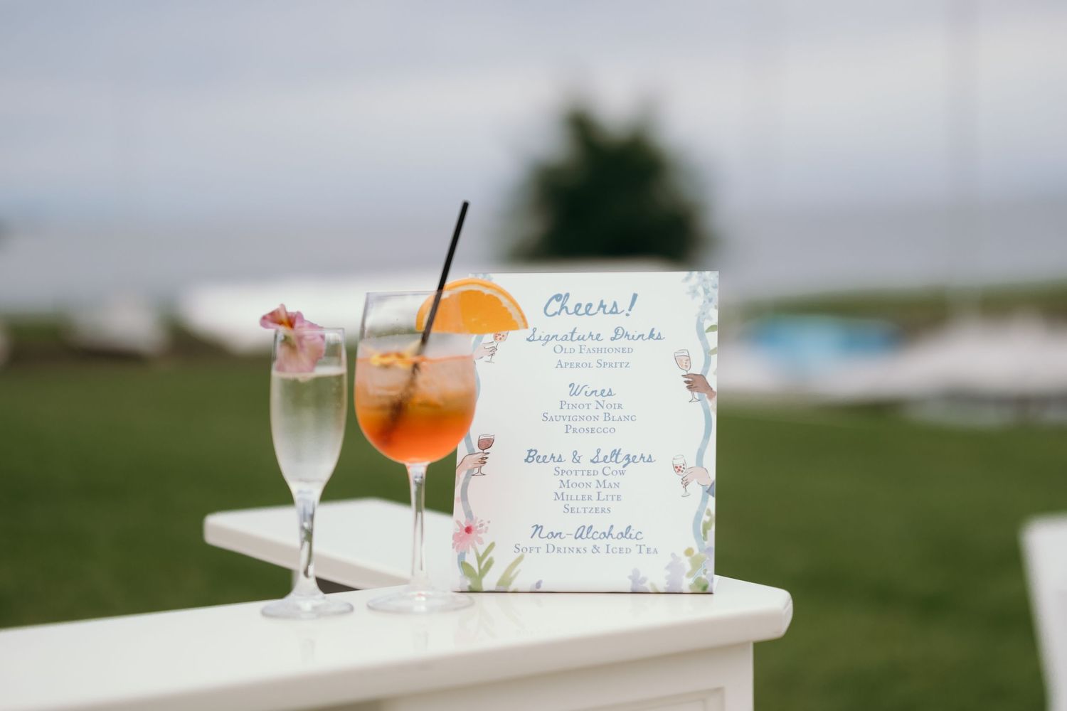 Two cocktails and a menu card sit on a white ledge overlooking a grassy outdoor venue.