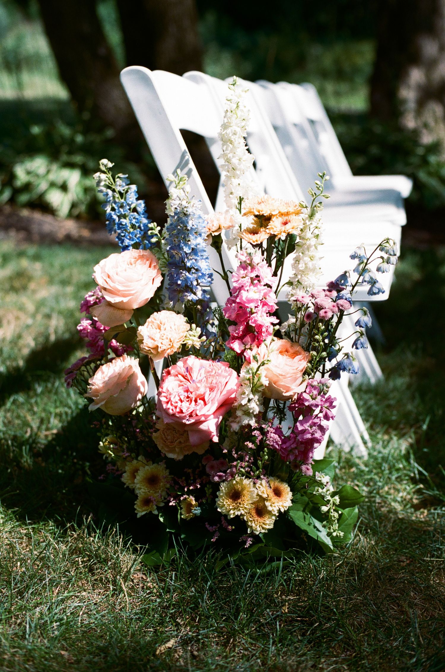 Lush outdoor wedding ceremony floral arrangement with pink roses, blue delphinium, and wildflowers beside white folding chairs on grass.