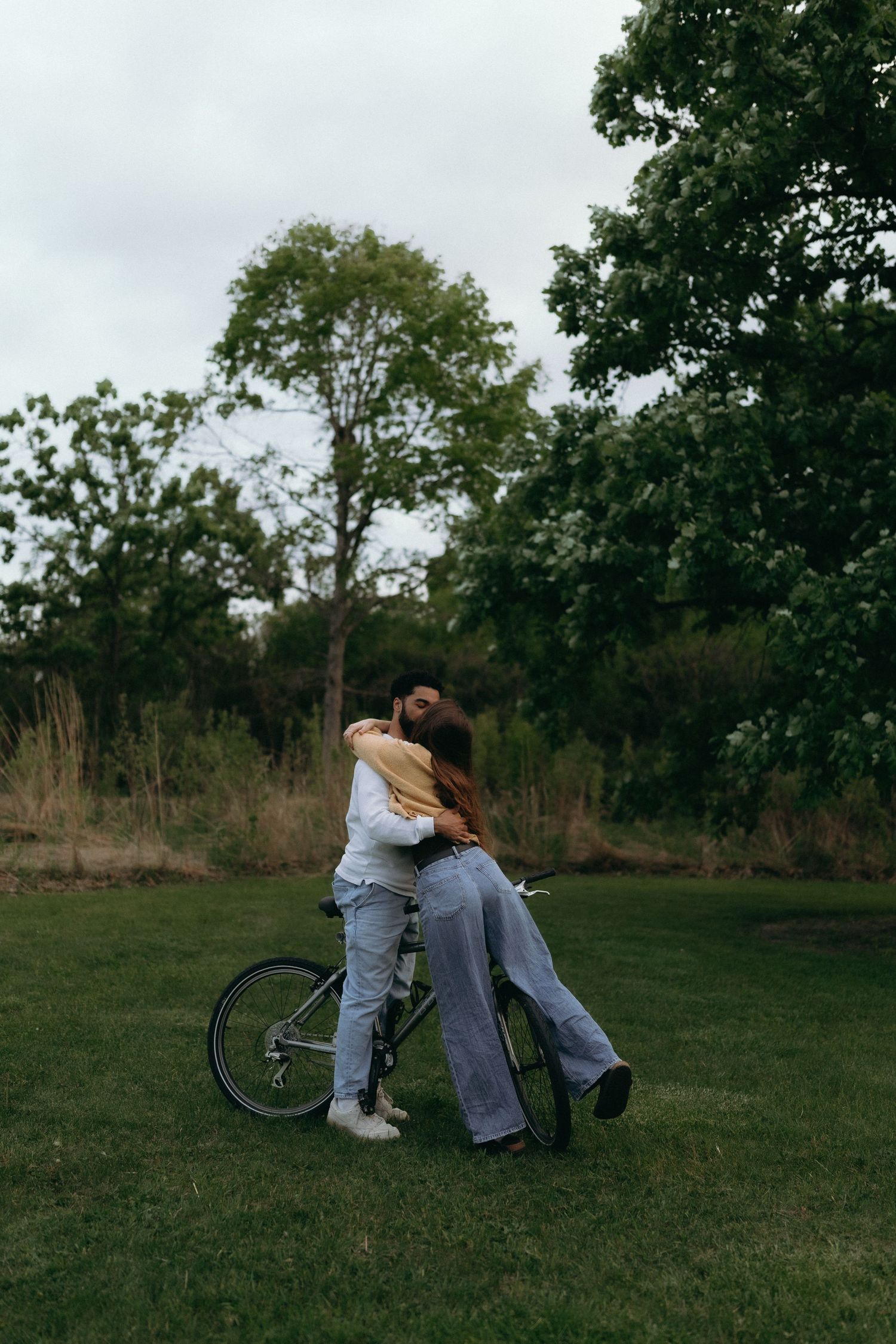 Chicago photogrpaher A couple embraces romantically in a green park with a bicycle beside them on a cloudy day.