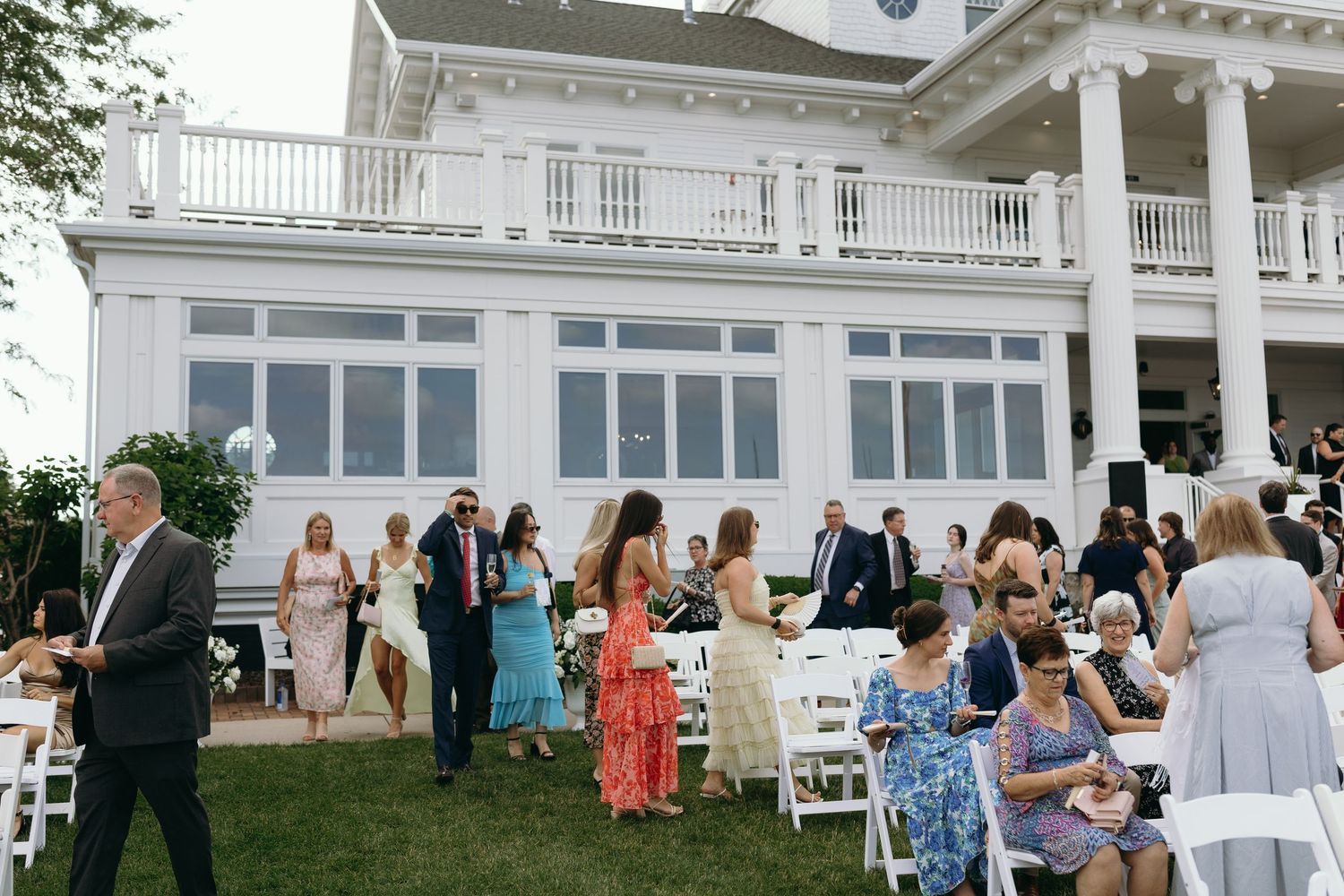 Wedding guests mingle and socialize outdoors at an elegant venue with white columns and balcony railings.