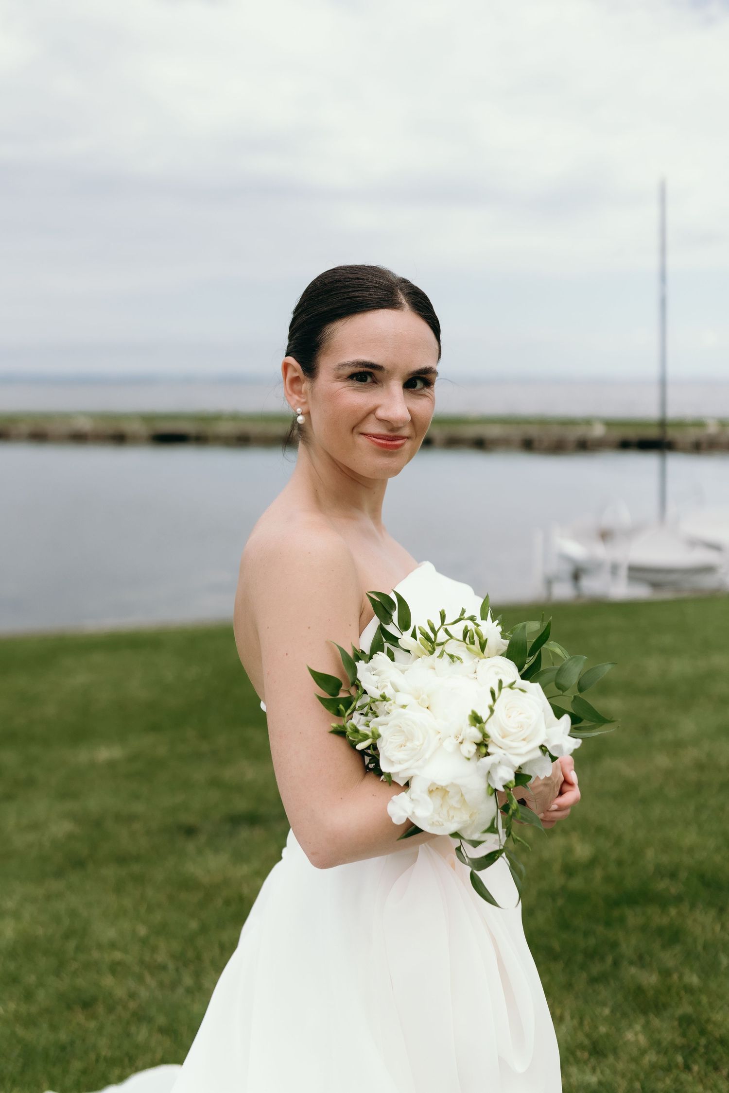A bride in a white strapless dress holds a stunning bouquet of white flowers and greenery by the waterfront. Oshosh wedding photographer