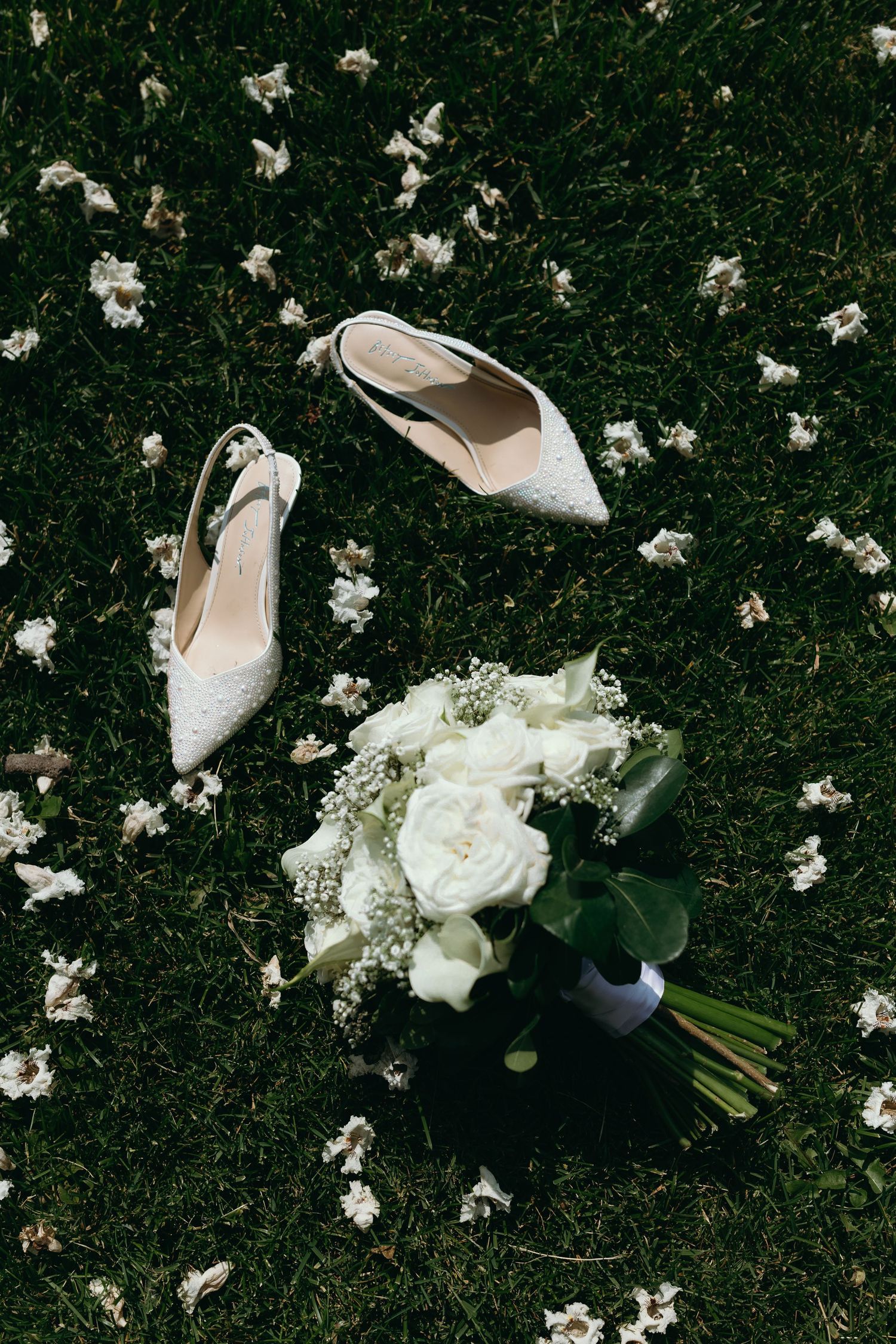White bridal shoes and bouquet arranged on grass with scattered flower petals.