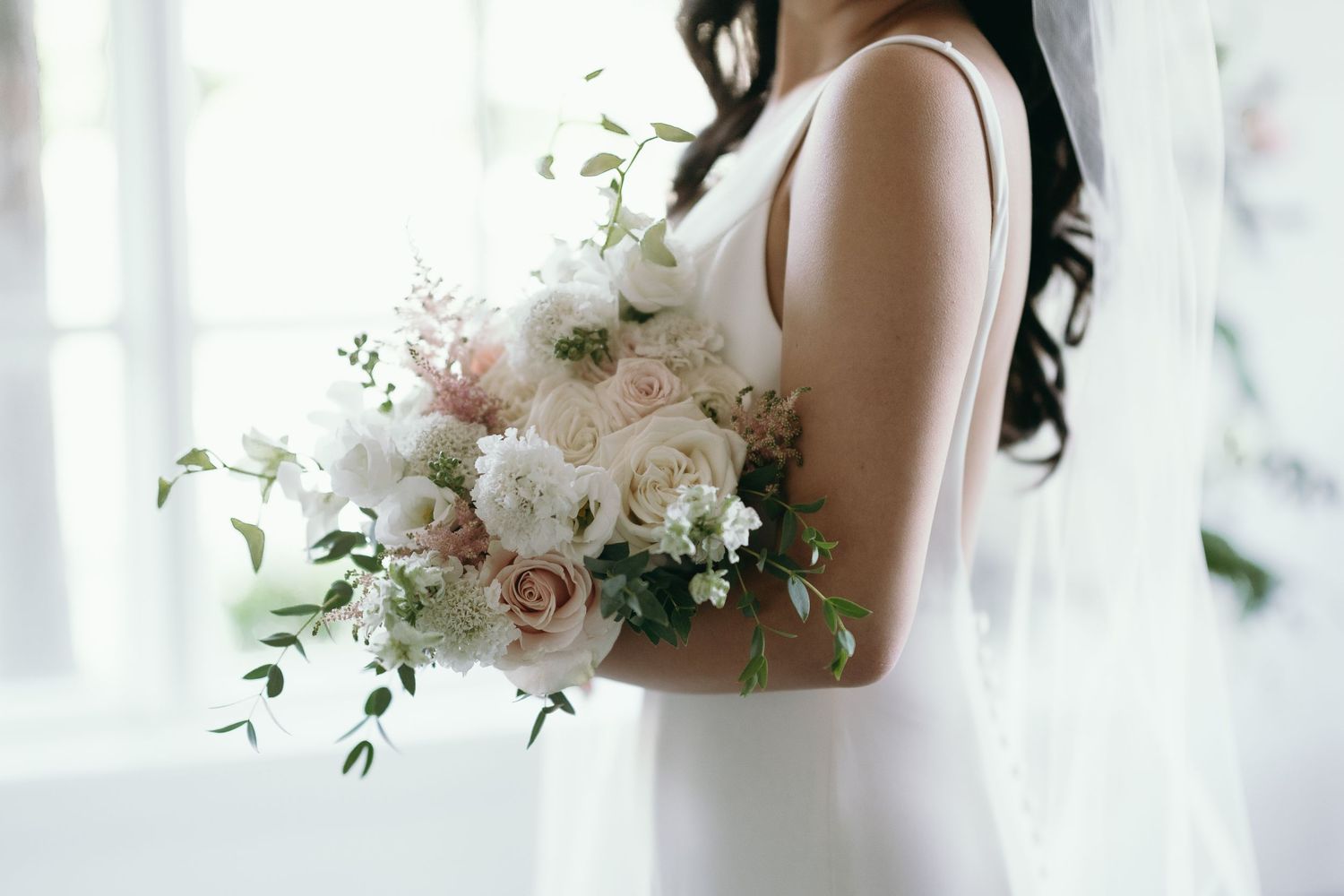 Side view of a delicate blush pink and white wedding bouquet with roses and greenery held against a light background.