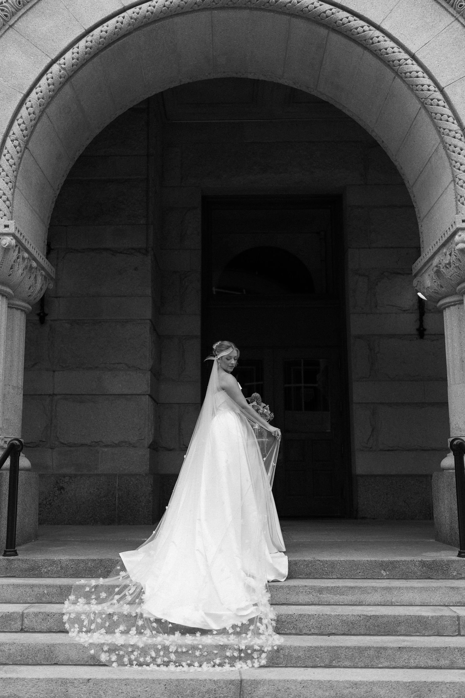 A bride in a flowing white wedding gown with a long train poses under a series of stone archways in black and white.