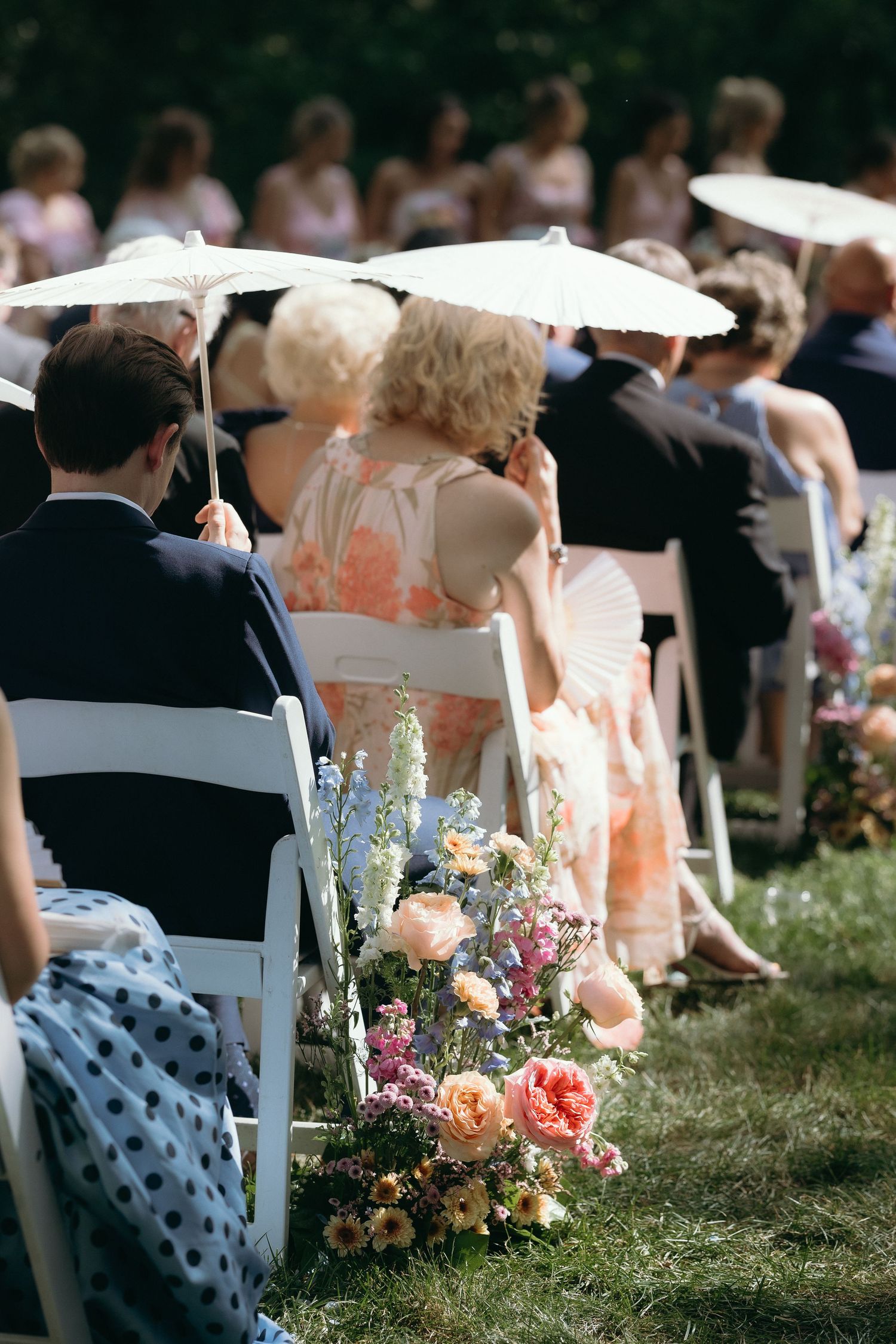 Outdoor wedding ceremony guests seated in white chairs along a floral aisle, holding parasols in summer sunshine.