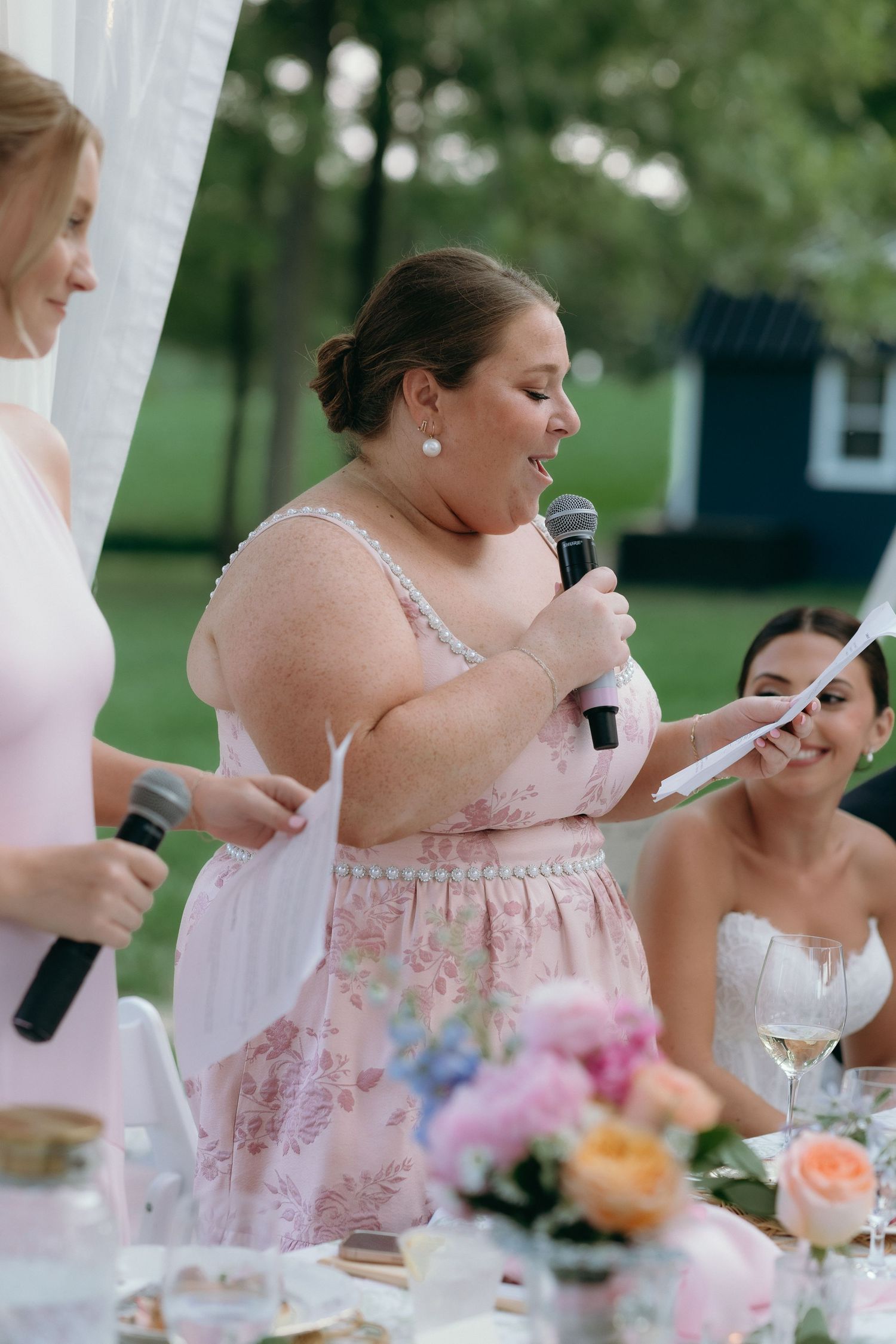 Bridesmaid in pink floral dress giving a heartfelt speech with microphone at outdoor wedding reception.