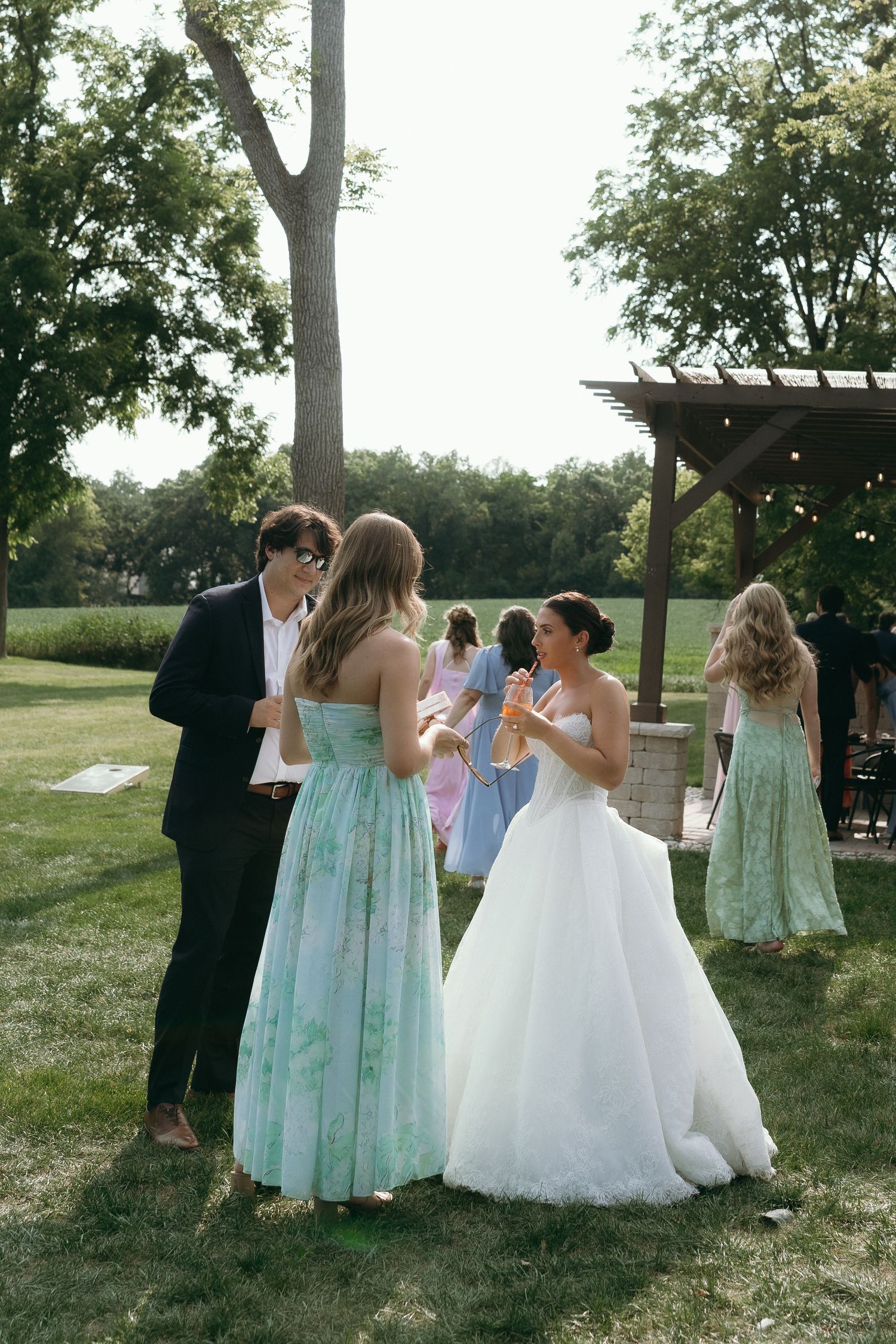 Bride in white gown chats with guests in floral dresses at an outdoor wedding reception on green lawn.
