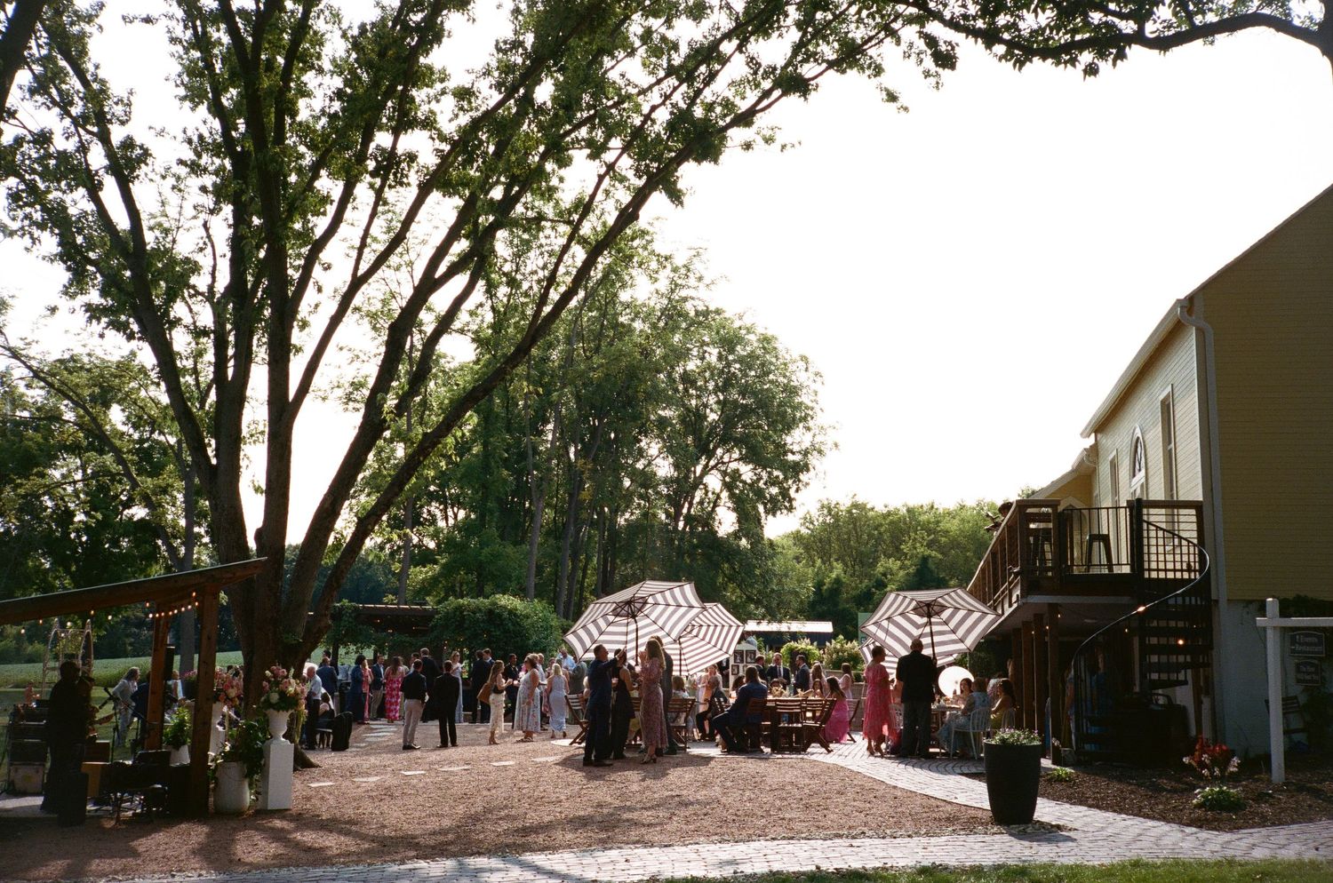 Outdoor wedding reception on a brick patio with striped umbrellas, lush trees, and guests mingling beside a building.