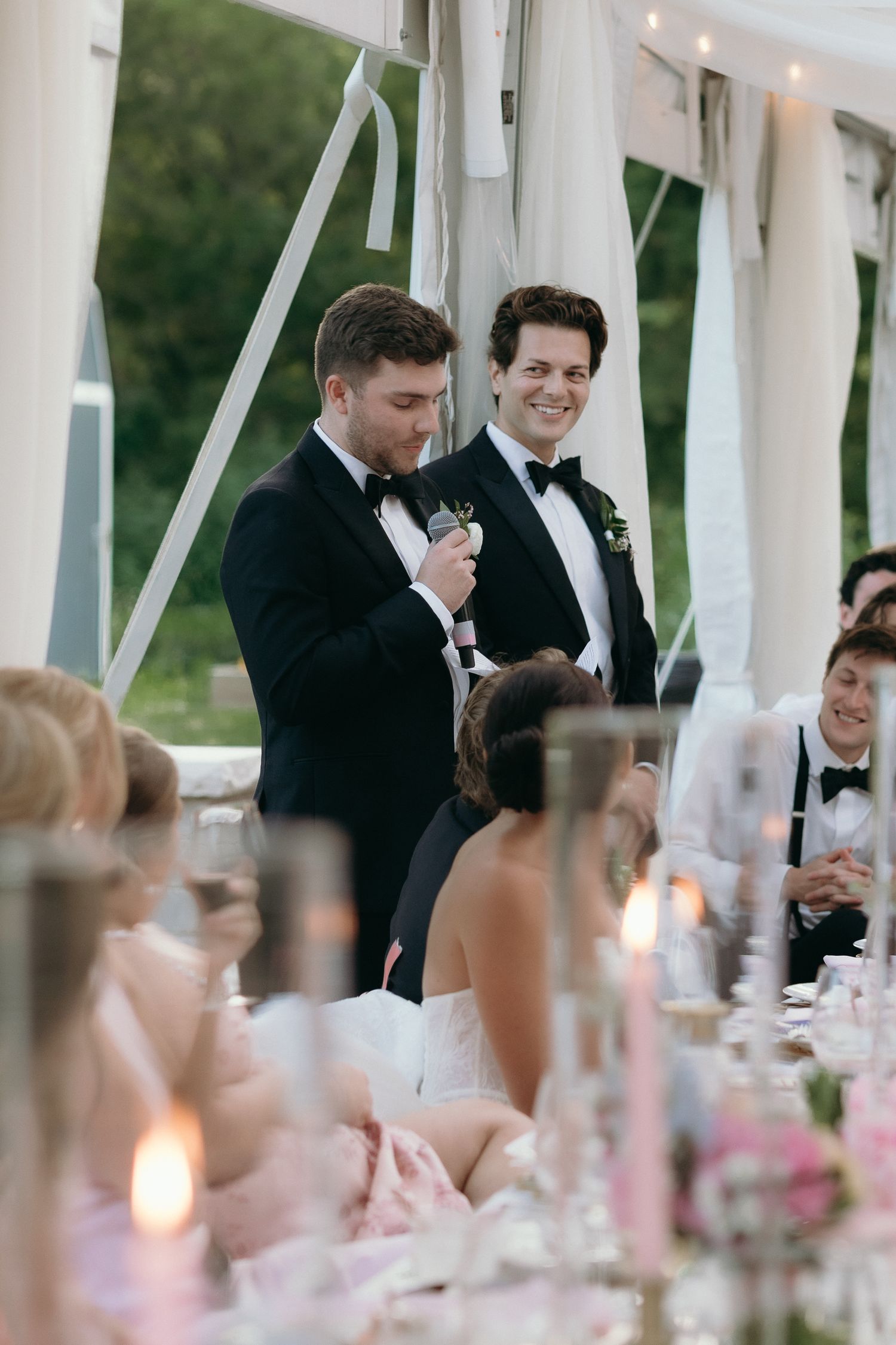 Two groomsmen in tuxedos delivering a wedding toast with microphone to seated reception guests.