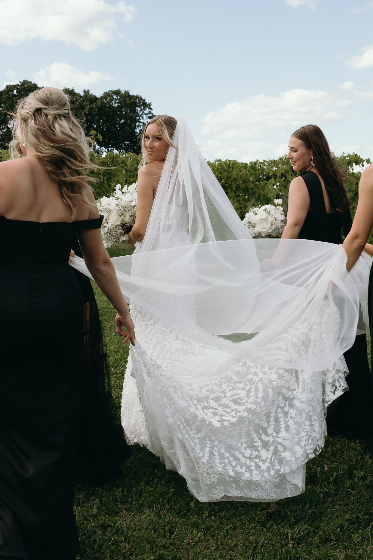 Bridesmaids in black dresses help arrange a flowing white lace wedding veil outdoors on a sunny day.