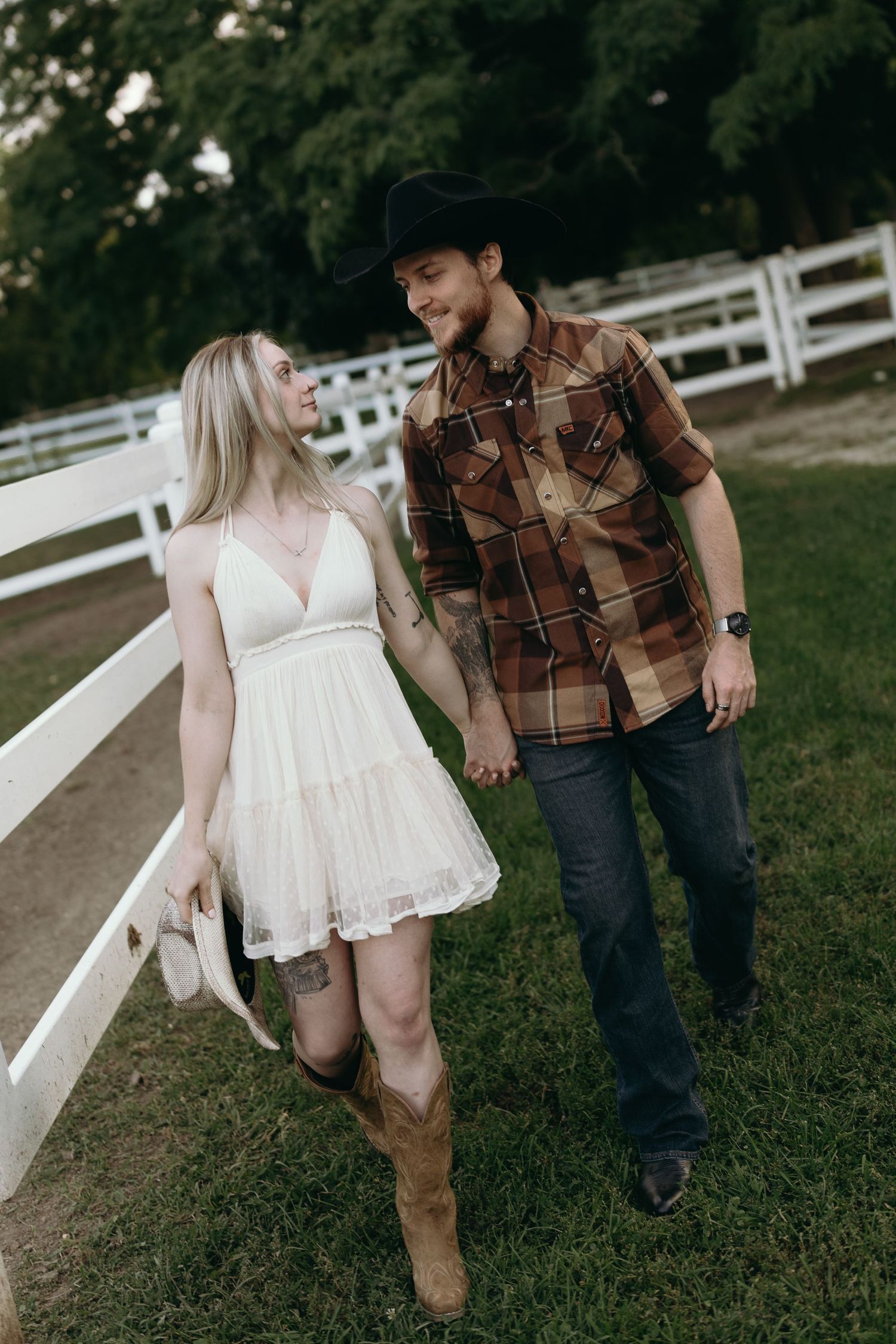 A couple in rustic attire walking together on a path by white fencing during an outdoor countryside photoshoot.